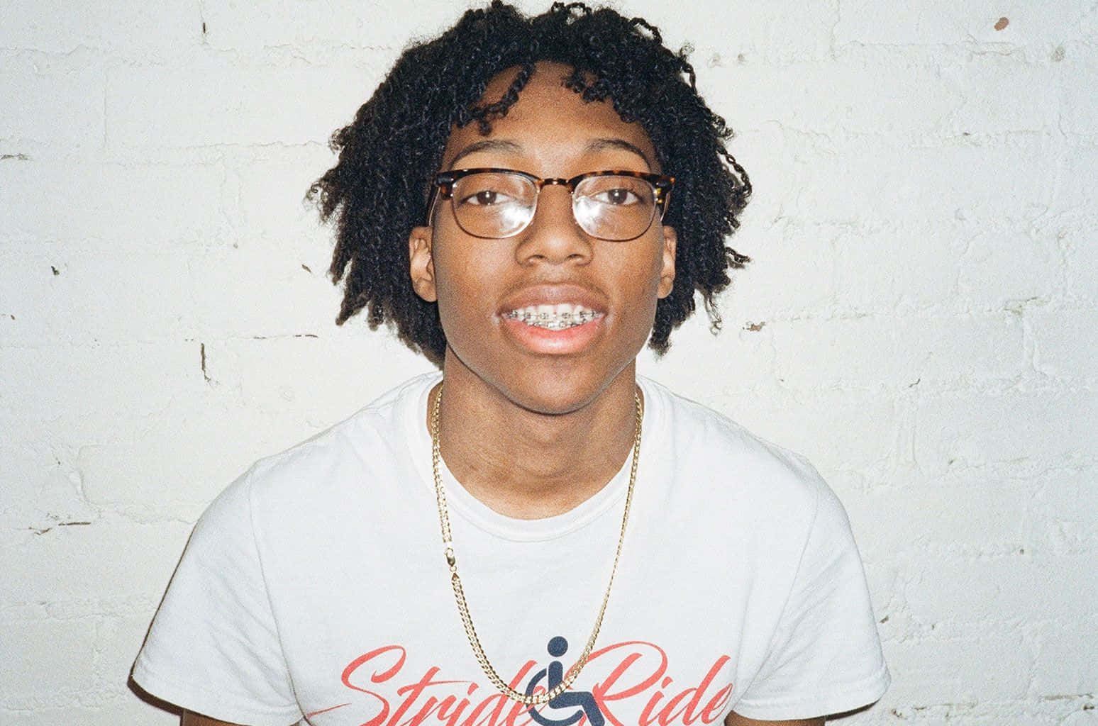 Rappers 桌布 壁紙 — 一位年輕人有捲髮和眼鏡，穿著白色T恤和金色鏈條，在白色磚牆前微笑。
