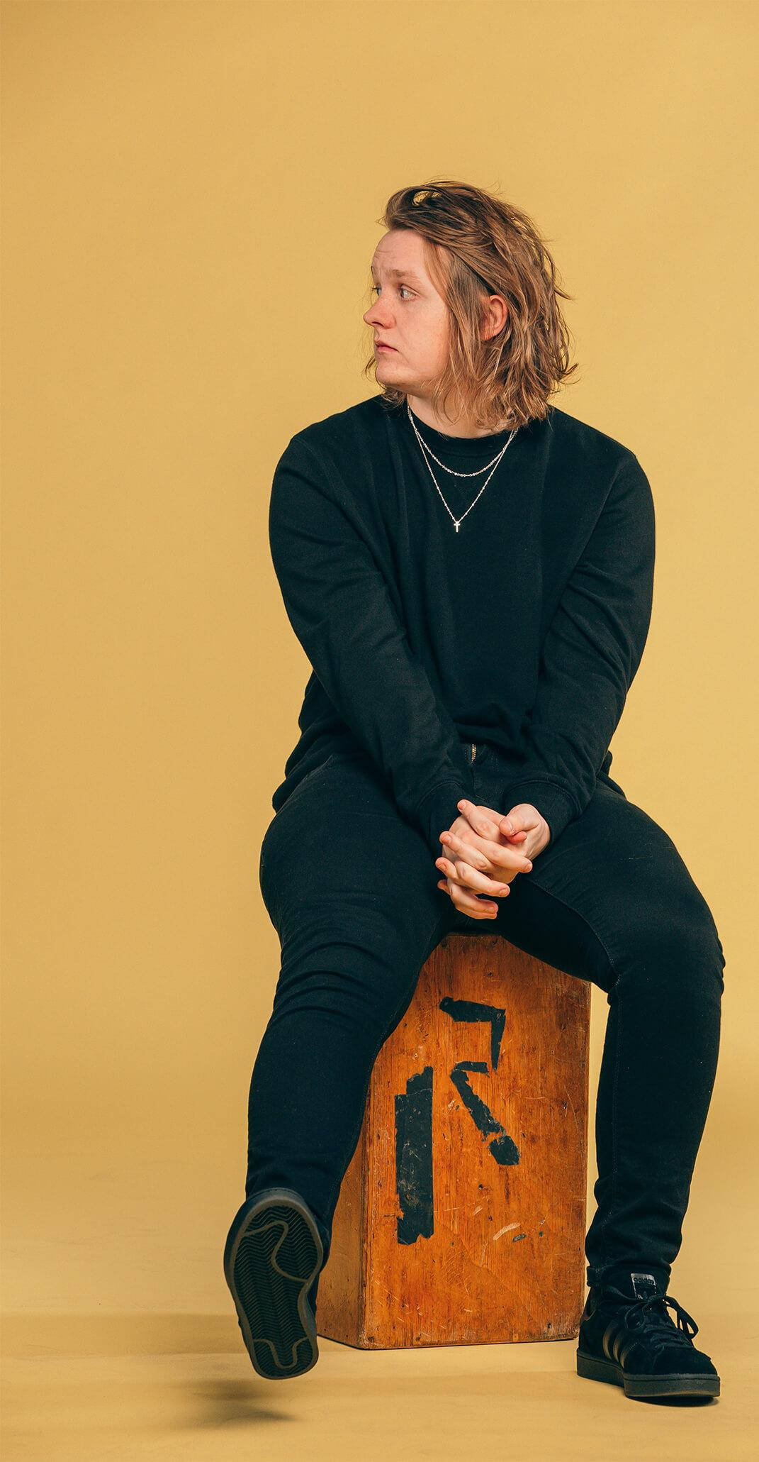 Lewis Capaldi 桌布 壁紙 — 一位年輕人有波浪頭髮，沉思地坐在木箱上，背景是溫暖的黃色攝影棚。