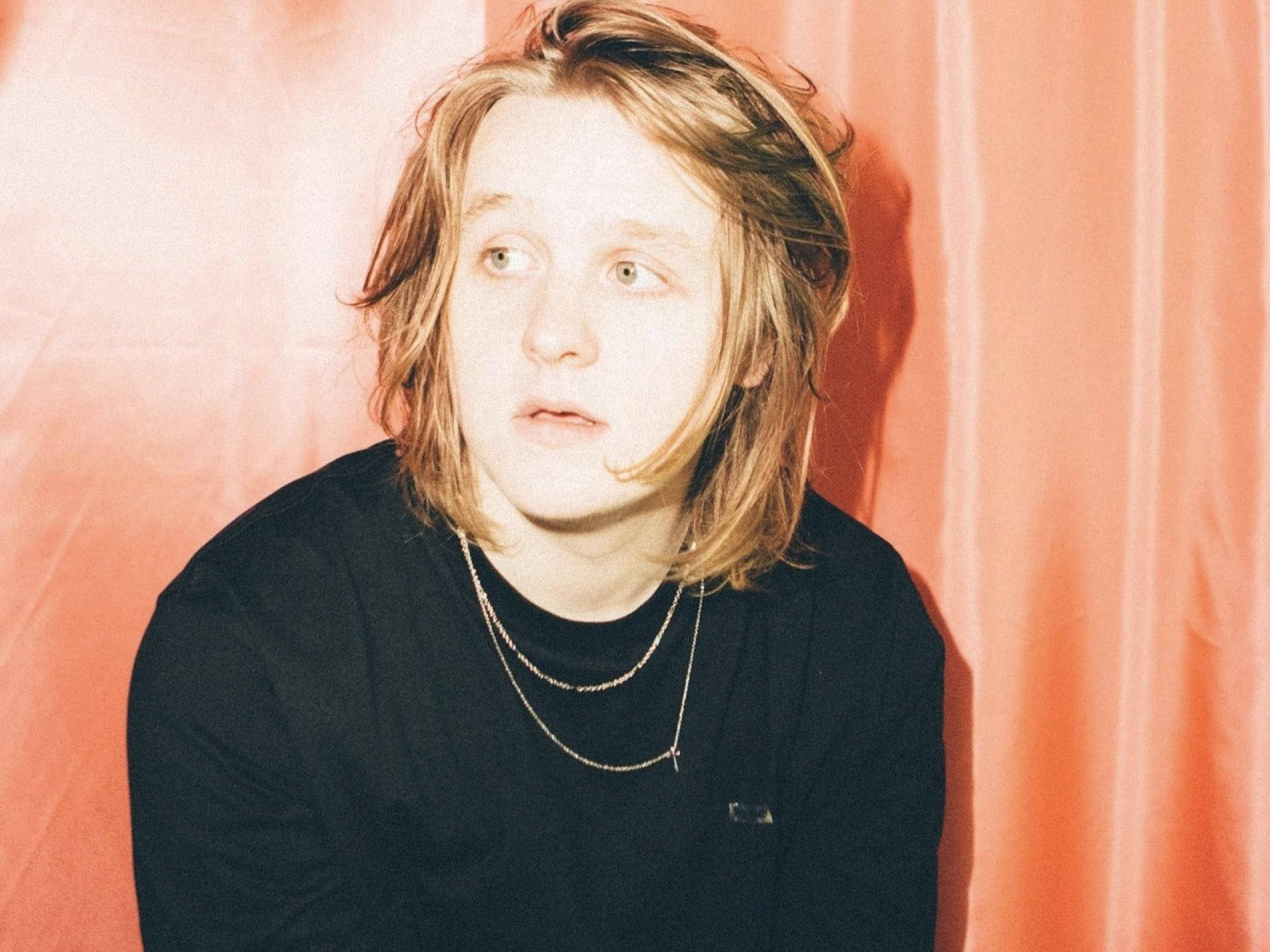 Lewis Capaldi 桌布 壁紙 — 一位年輕人有肩長金髮，穿著黑色襯衫和層疊項鍊，背景是柔和的珊瑚色窗簾。