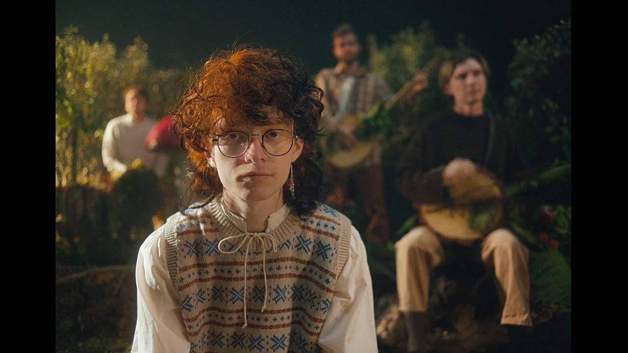 Cavetown 桌布 壁紙 — 一位年輕人有著捲曲的紅髮和眼鏡，在溫暖的傍晚光線中凝視前方，身後有音樂家在花園中演奏。