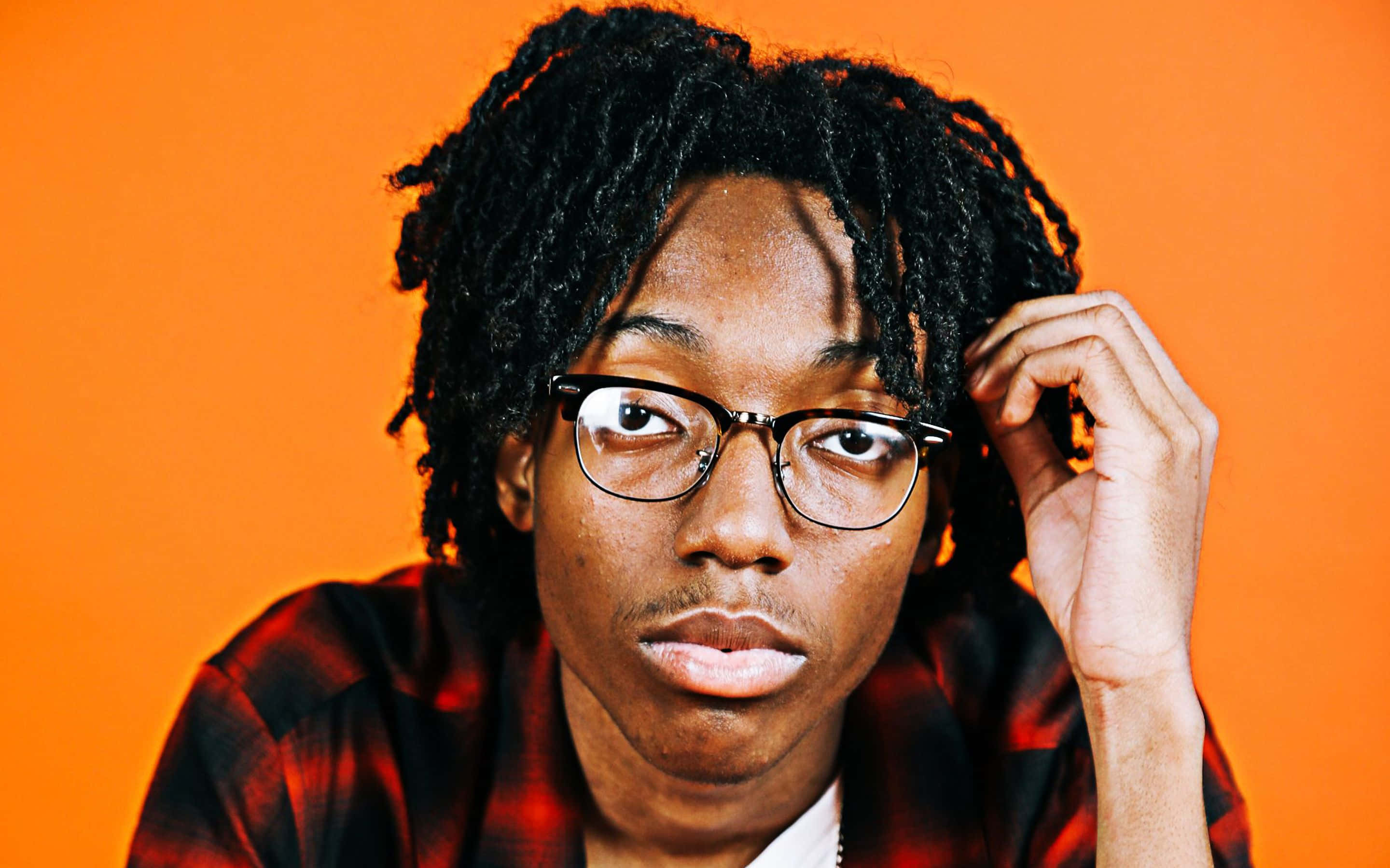 Rappers 桌布 壁紙 — 一位年輕人有辮子頭和眼鏡，在充滿活力的橙色背景下若有所思地凝視。