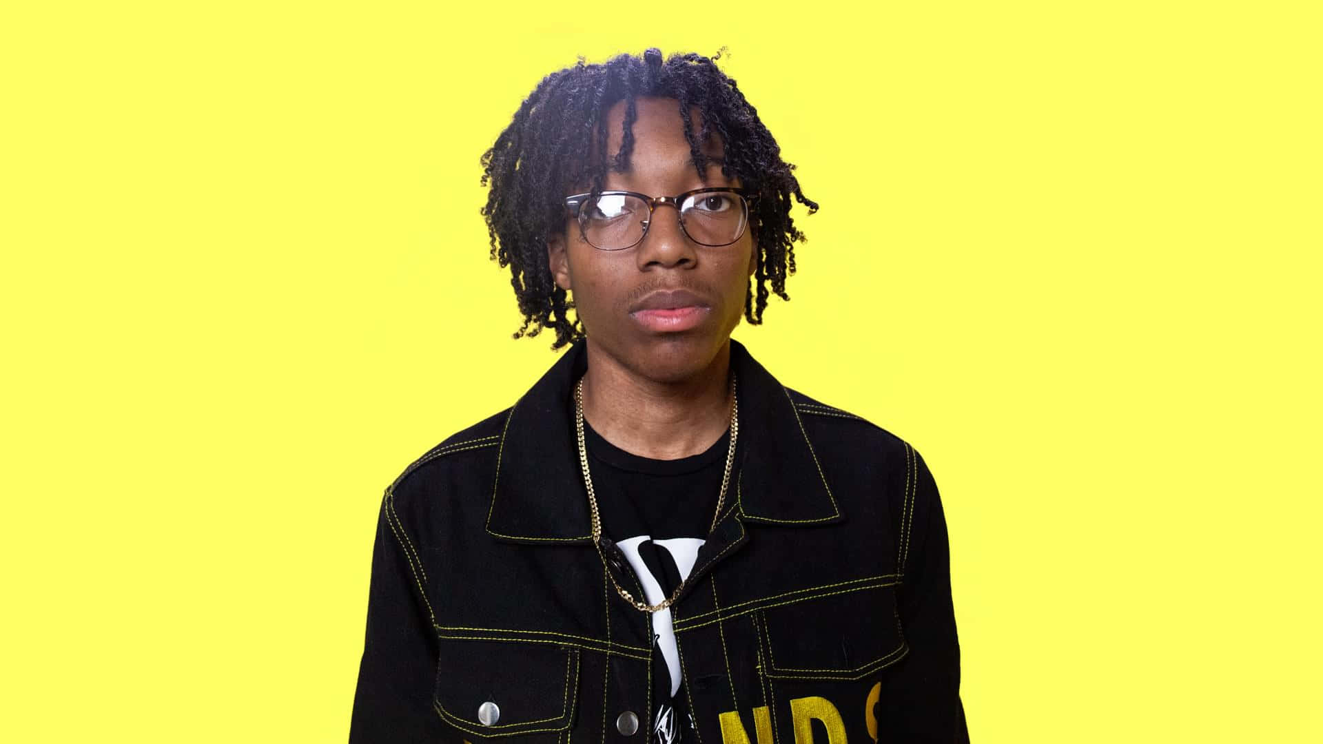 Rappers 桌布 壁紙 — 一位年輕人有辮子頭和眼鏡，在明亮的黃色背景下直視鏡頭。