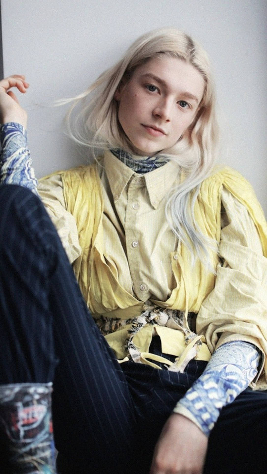 Hunter Schafer 桌布 壁紙 — 一位年輕人有鉑金色頭髮，隨意地靠著素牆坐著，穿著分層黃色襯衫和圖案袖子。