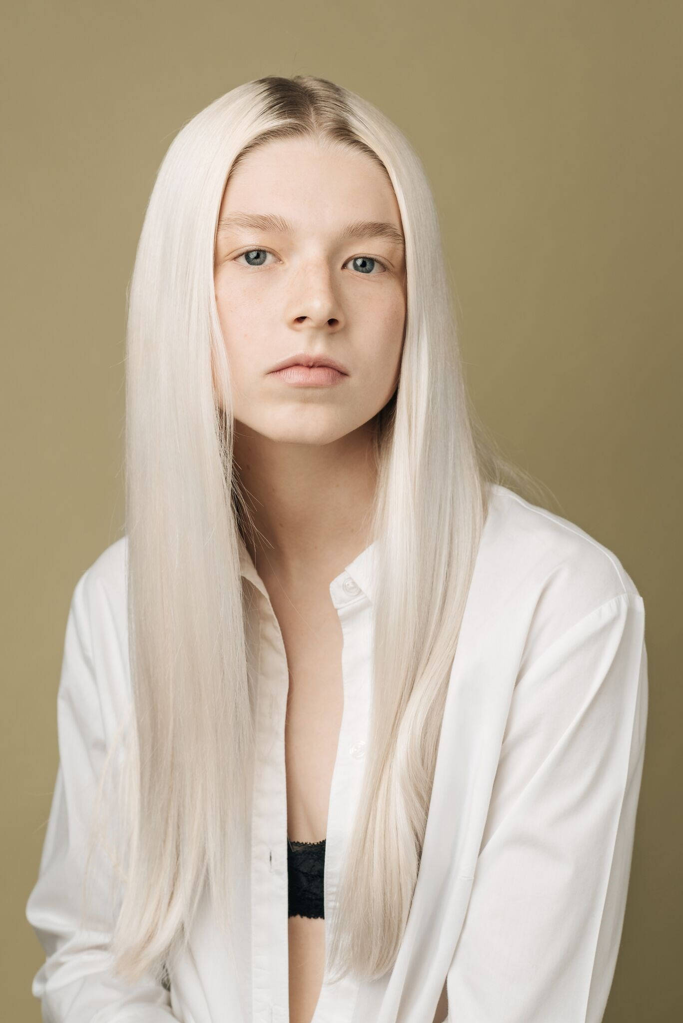 Hunter Schafer 桌布 壁紙 — 一位年輕人有長白金髮和藍眼睛，穿著敞開的白襯衫，在柔和的橄欖綠背景前直視鏡頭。