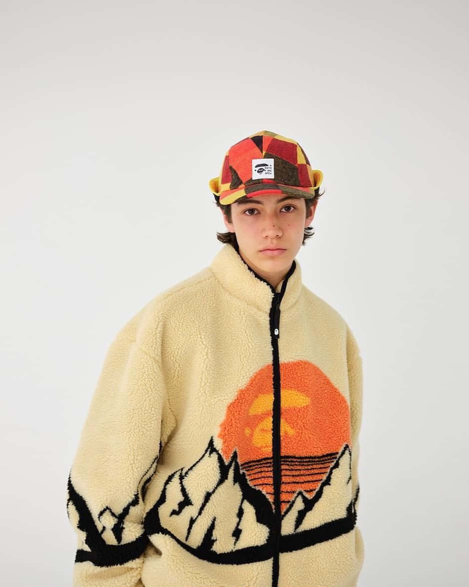 Bape 動態 桌布 壁紙 — 一位年輕人穿著奶油色絨毛外套，上面有醒目的山景日落圖案，戴著彩色拼布帽，背景為純白色。