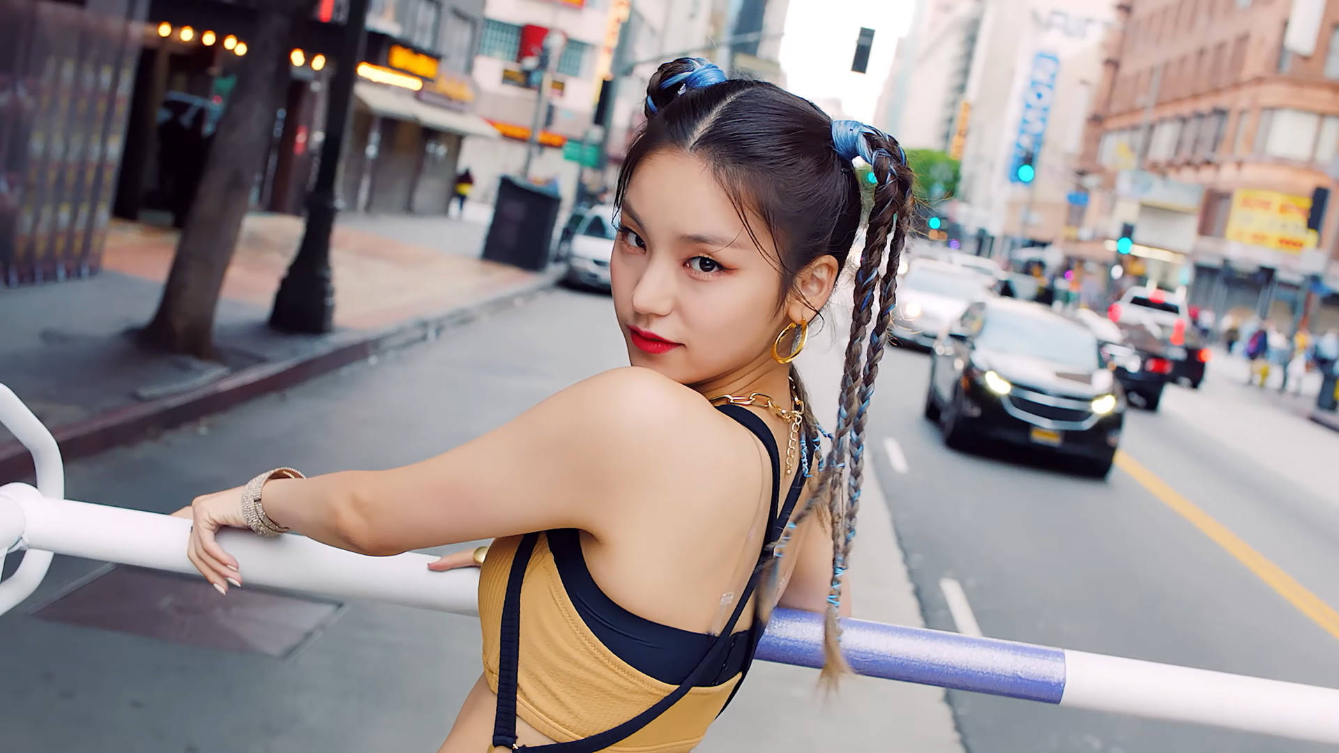 ITZY 桌布 壁紙 — 一位年輕女子，辮子末端染藍，倚靠在繁華城市景觀的街道欄杆上，回頭凝視，紅唇大膽，戴著金色耳環。