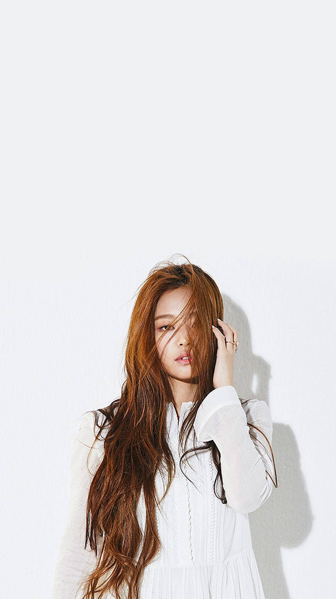 Jennie 桌布 壁紙 — 一位年輕女子，長著赤褐色長髮，身穿白色連衣裙，站在純白牆壁前，她的手輕輕觸摸臉龐，柔和的光線在她身後投下陰影。