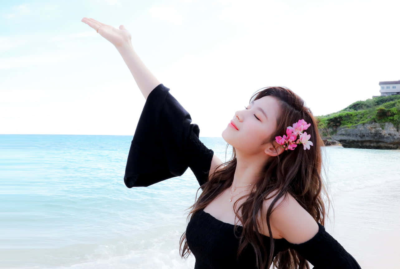 twice 桌布 壁紙 — 一位年輕女子，頭髮上插著粉紅色的花朵，在明亮的沙灘上，面對著碧綠的海浪，將手臂伸向天空。