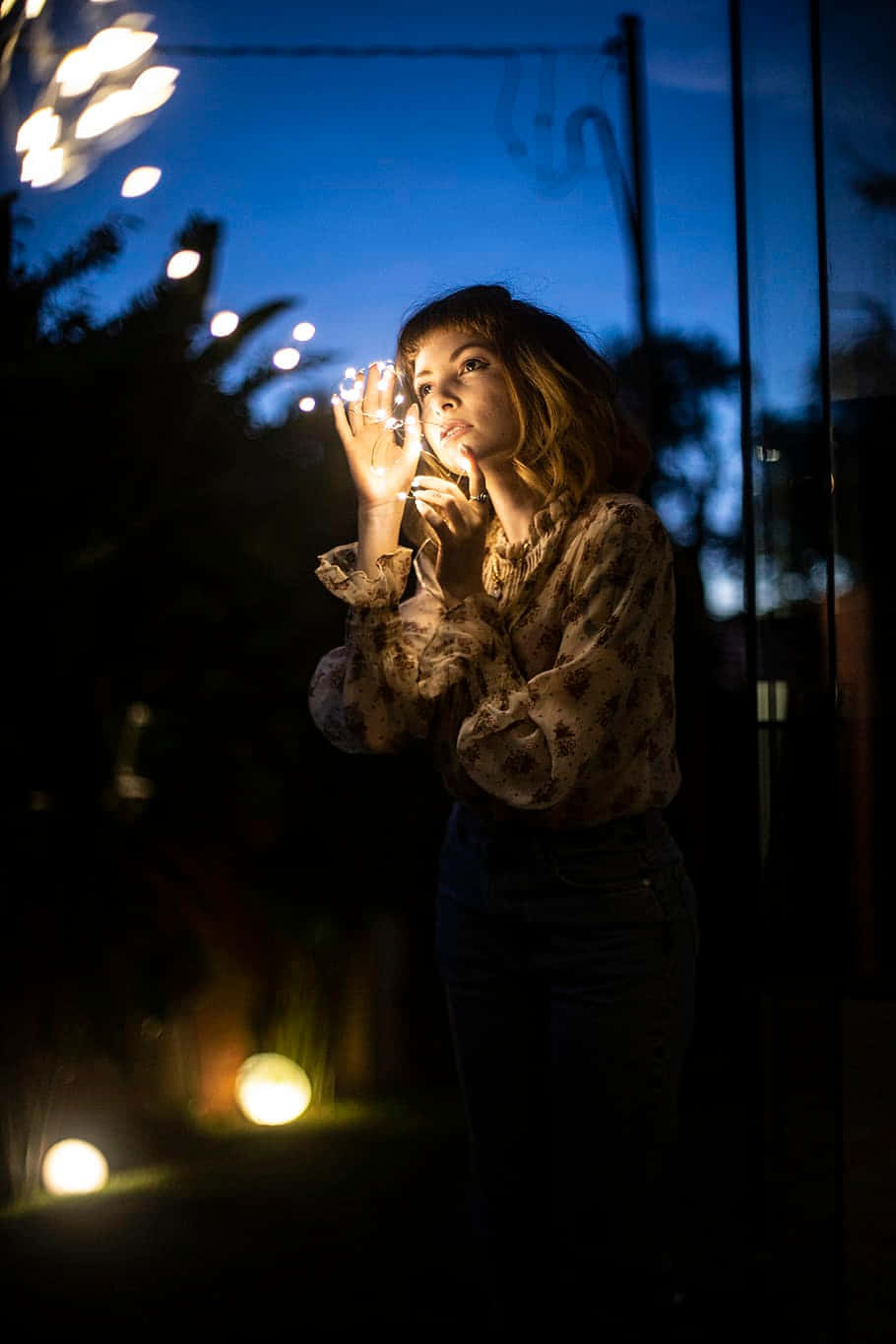 Light Bulb 桌布 壁紙 — 一位年輕女子在夜晚站立，她的雙手在深藍色天空和模糊的溫暖燈籠襯托下發出柔和的光芒。