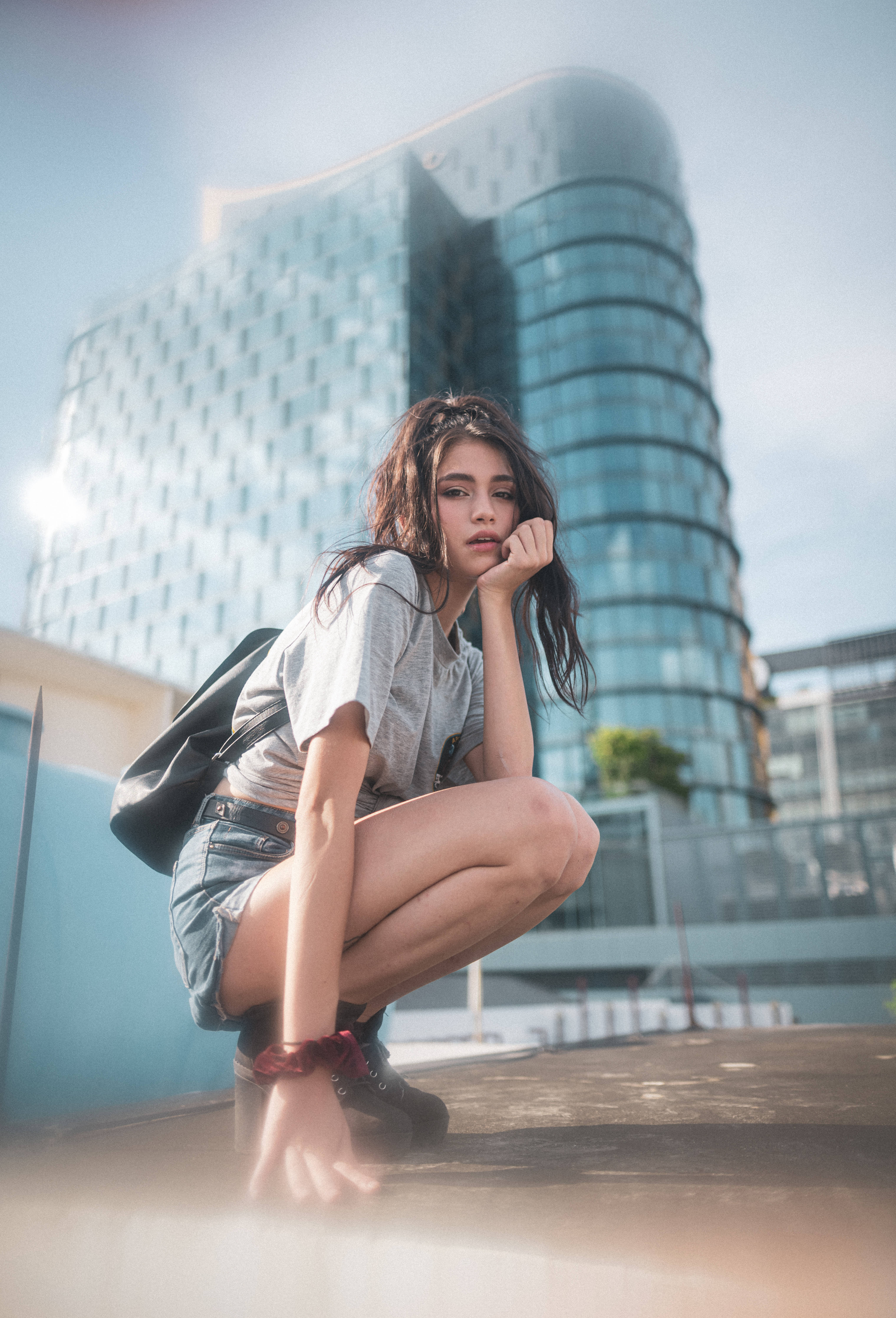 Teenage 桌布 壁紙 — 一位年輕女子在屋頂上若有所思地蹲著，背後是一座現代玻璃摩天大樓在明亮的日光下高聳入雲。