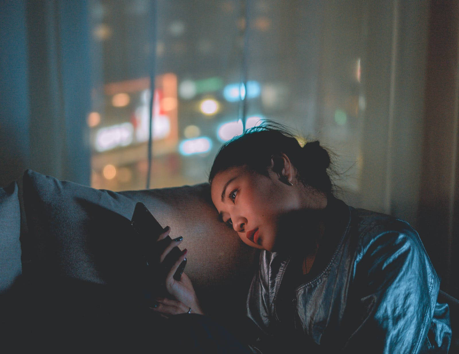 Lonely 桌布 壁紙 — 一位年輕女子將頭靠在沙發枕頭上，在身後窗戶可見的城市燈光下，專注地看著她的手機。