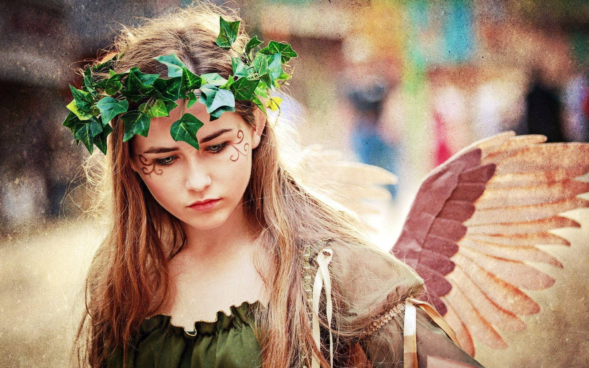 witchcraft 桌布 壁紙 — 一位年輕女子戴著常春藤冠冕和棕色翅膀，在柔和光線、夢幻般的戶外場景中向下凝視。