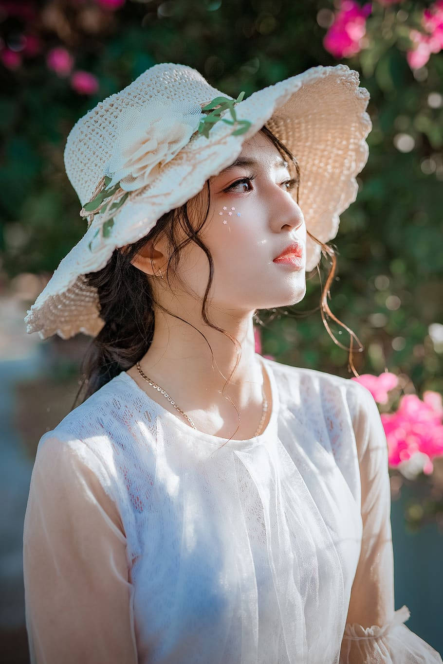 女孩 桌布 壁紙 — 一位年輕女子戴著白色編織帽，身穿透視襯衫，在柔和的粉紅花朵和斑駁的陽光中向上凝視。