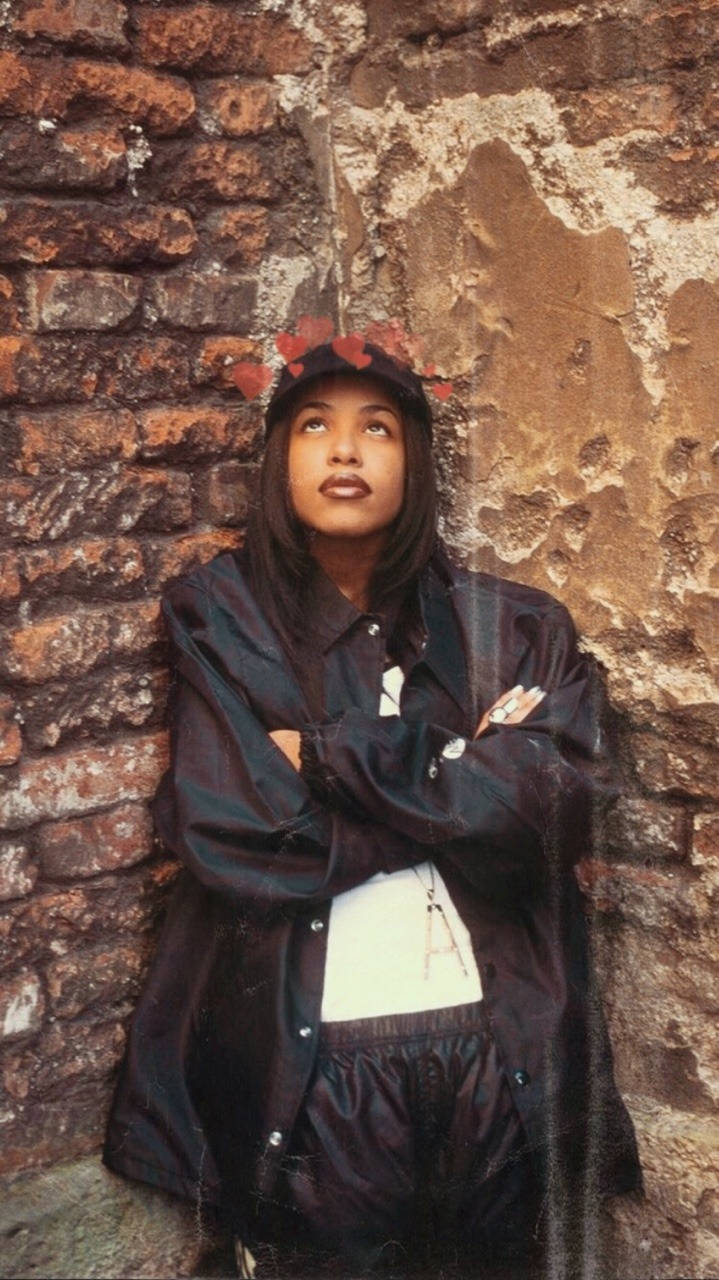 aaliyah 桌布 壁紙 — 一位年輕女子戴著紅色花冠，靠在一面風化的磚牆上，雙臂交叉，抬頭凝視，表情若有所思。