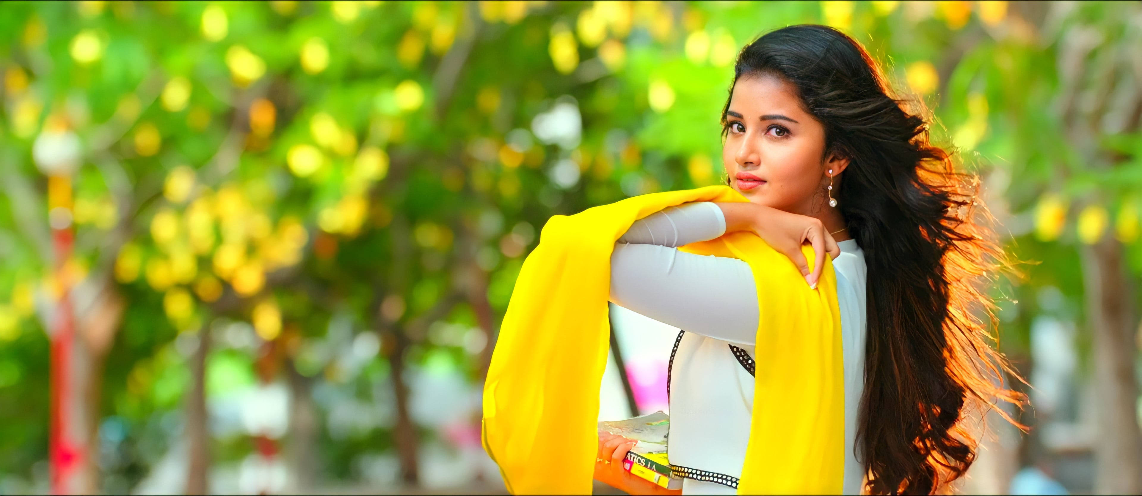 anupama parameswaran 桌布 壁紙 — 一位年輕女子披著飄逸的深色長髮，圍著鮮黃色圍巾，在陽光普照的綠葉背景下優雅地擺姿勢。