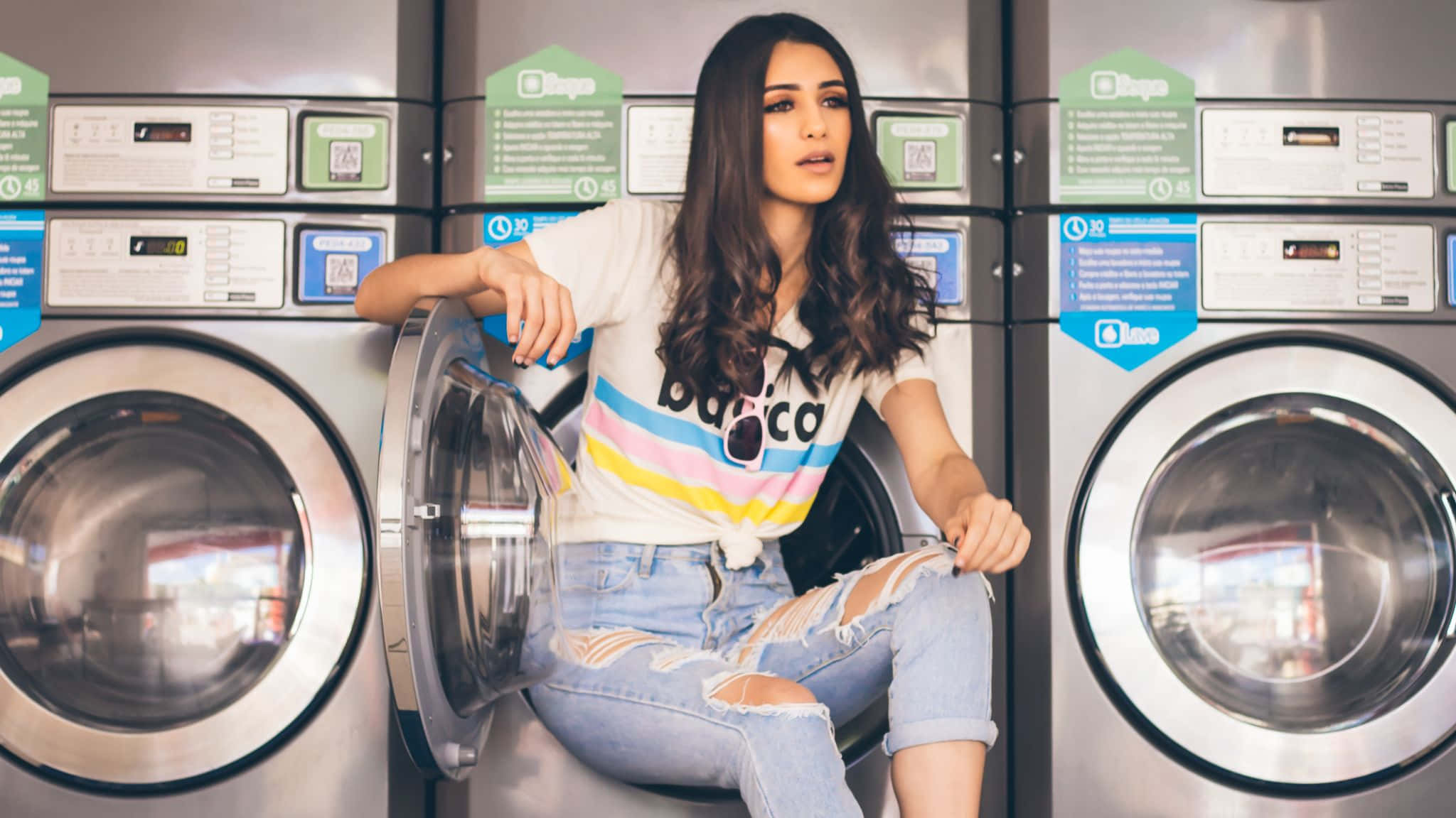 Laundry Room 桌布 壁紙 — 一位年輕女子留著深色長髮，隨意地坐在洗衣店的前置式洗衣機上，穿著彩色條紋T恤和破洞牛仔褲。