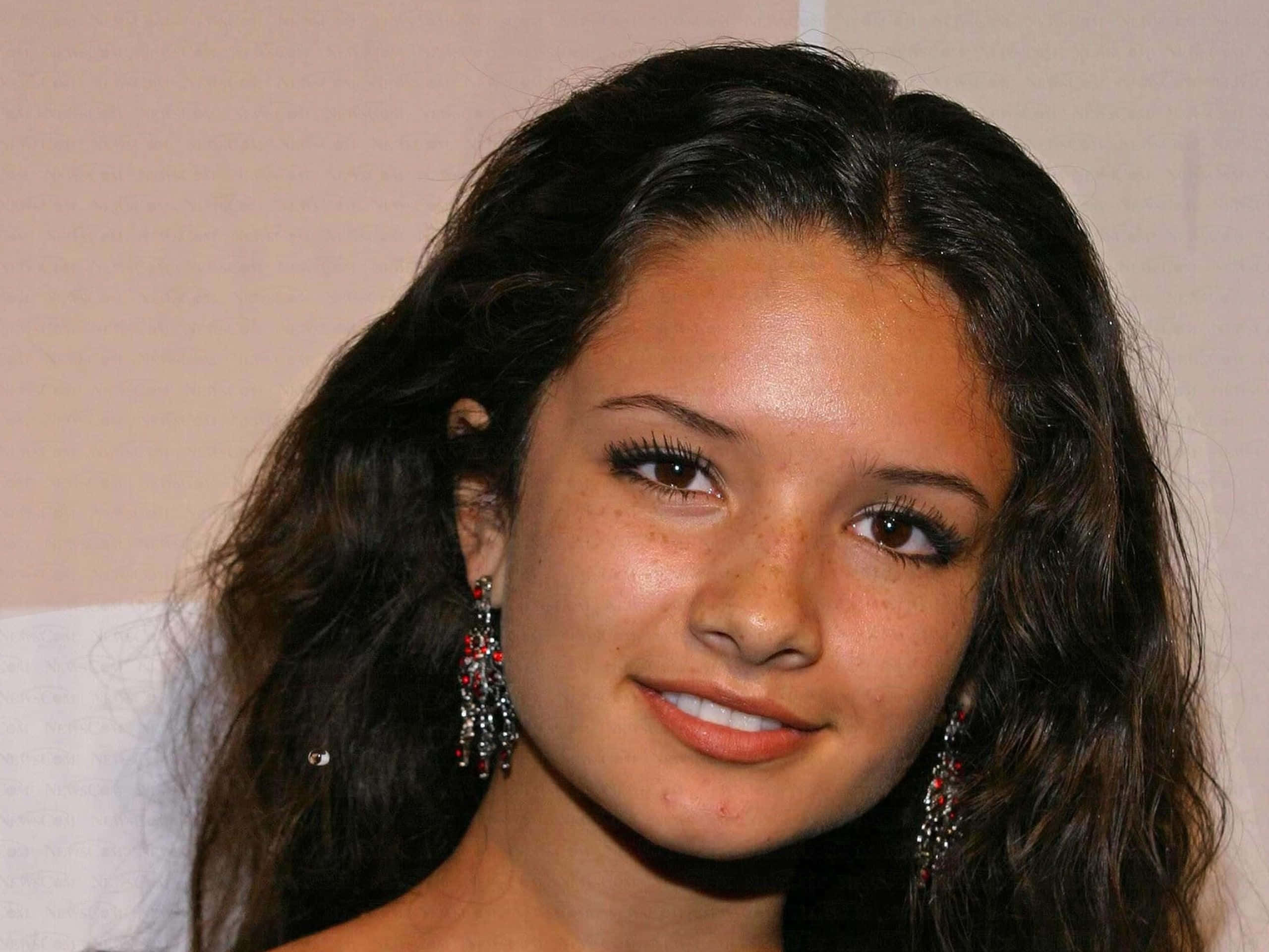 Alice Greczyn 桌布 壁紙 — 一位年輕女子的特寫肖像，她擁有深色波浪髮和雀斑，戴著閃亮的紅色和銀色耳環，溫柔地微笑。