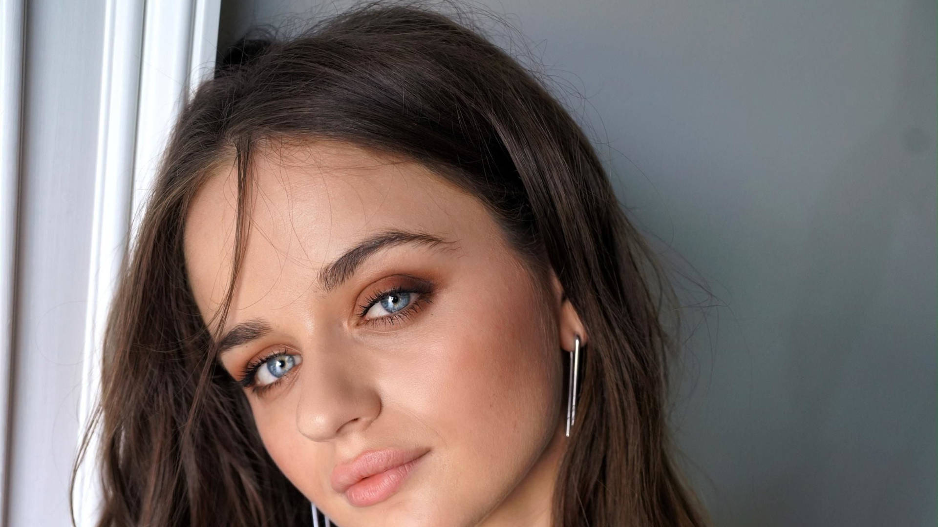 Joey King 桌布 壁紙 — 一位年輕女子的特寫肖像，她有引人注目的藍眼睛和深色頭髮，靠在窗框上。
