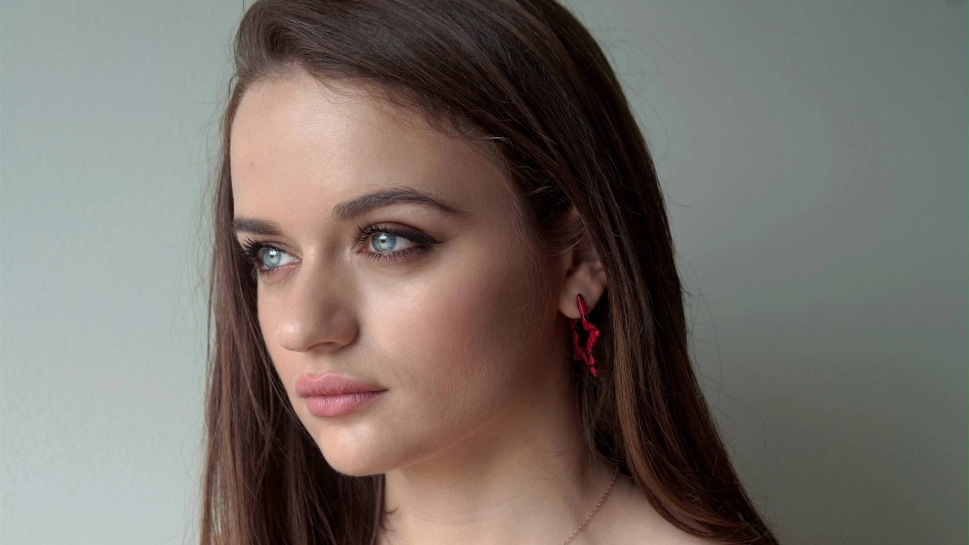 Joey King 桌布 壁紙 — 一位年輕女子的特寫肖像，擁有引人注目的藍色眼睛和深色頭髮，戴著紅色星星耳環，背景是柔和的灰色。