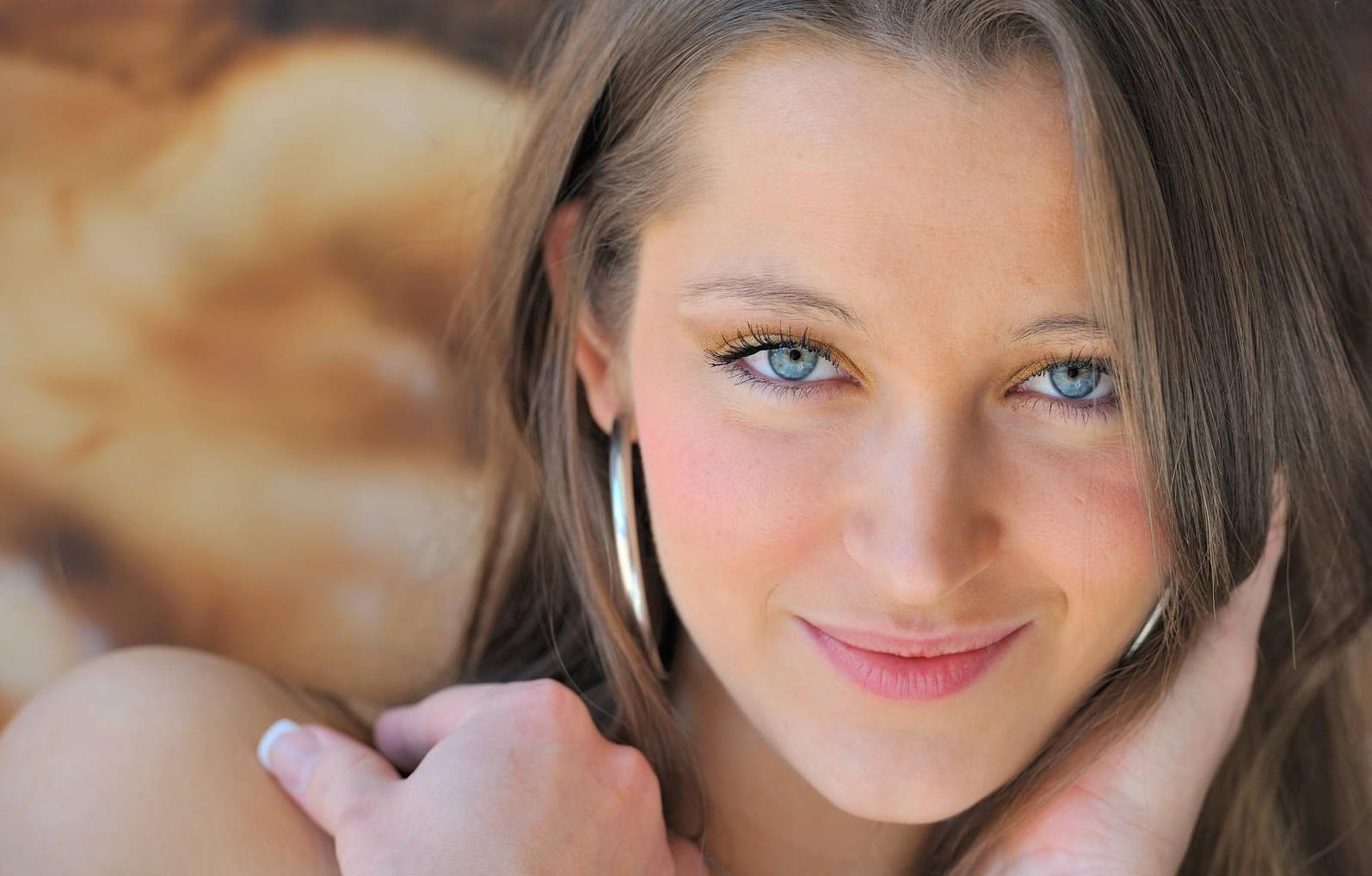 Dani Daniels 桌布 壁紙 — 一位年輕女子的特寫肖像，擁有引人注目的藍色眼睛和棕色頭髮，輕輕微笑，下巴擱在手上。