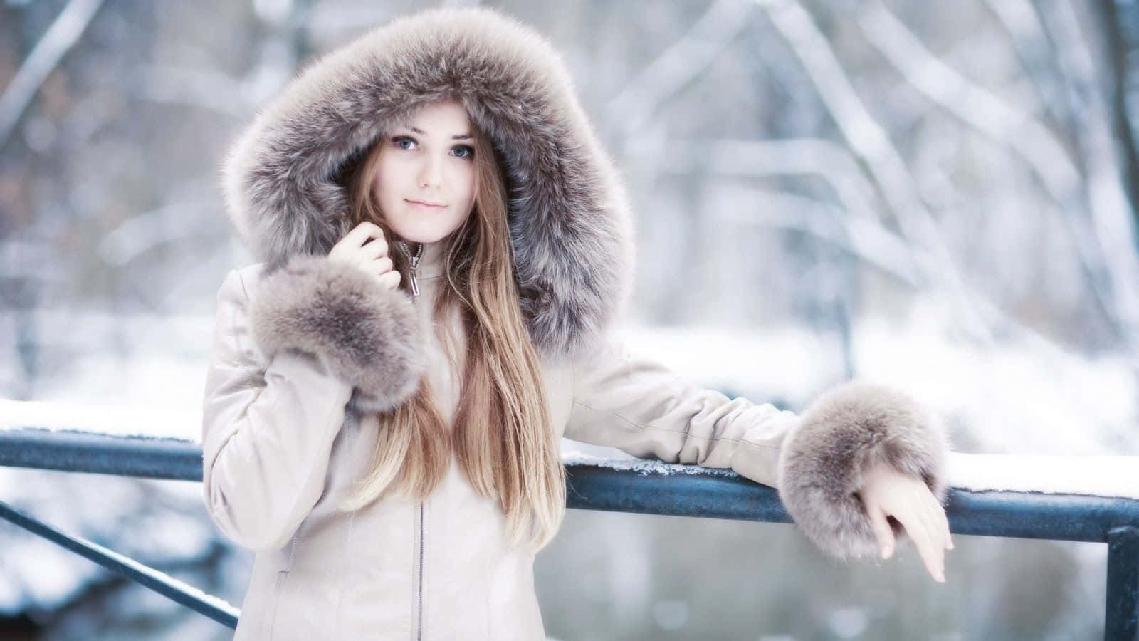 Winter Fashion 桌布 壁紙 — 一位年輕女子穿著毛領冬季外套，站在積雪的欄杆旁，身後是光禿禿的樹木。