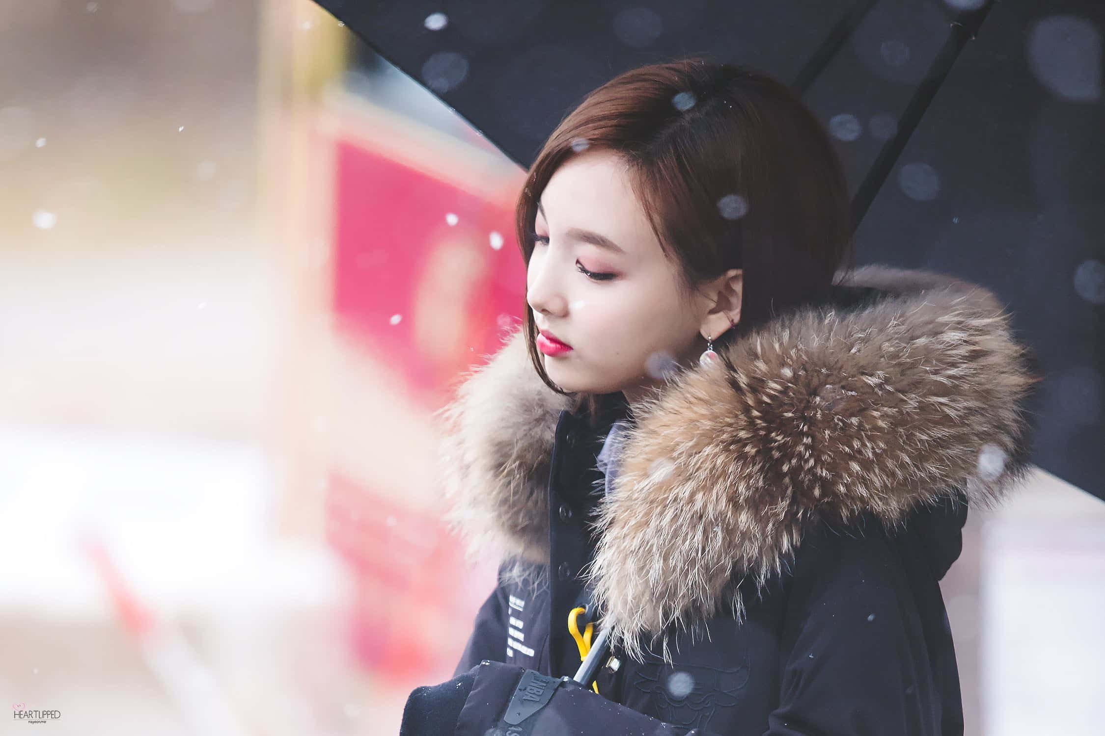 twice 桌布 壁紙 — 一位年輕女子穿著毛領外套，在雪花飄落的柔和模糊城市背景下，撐著一把黑色雨傘。