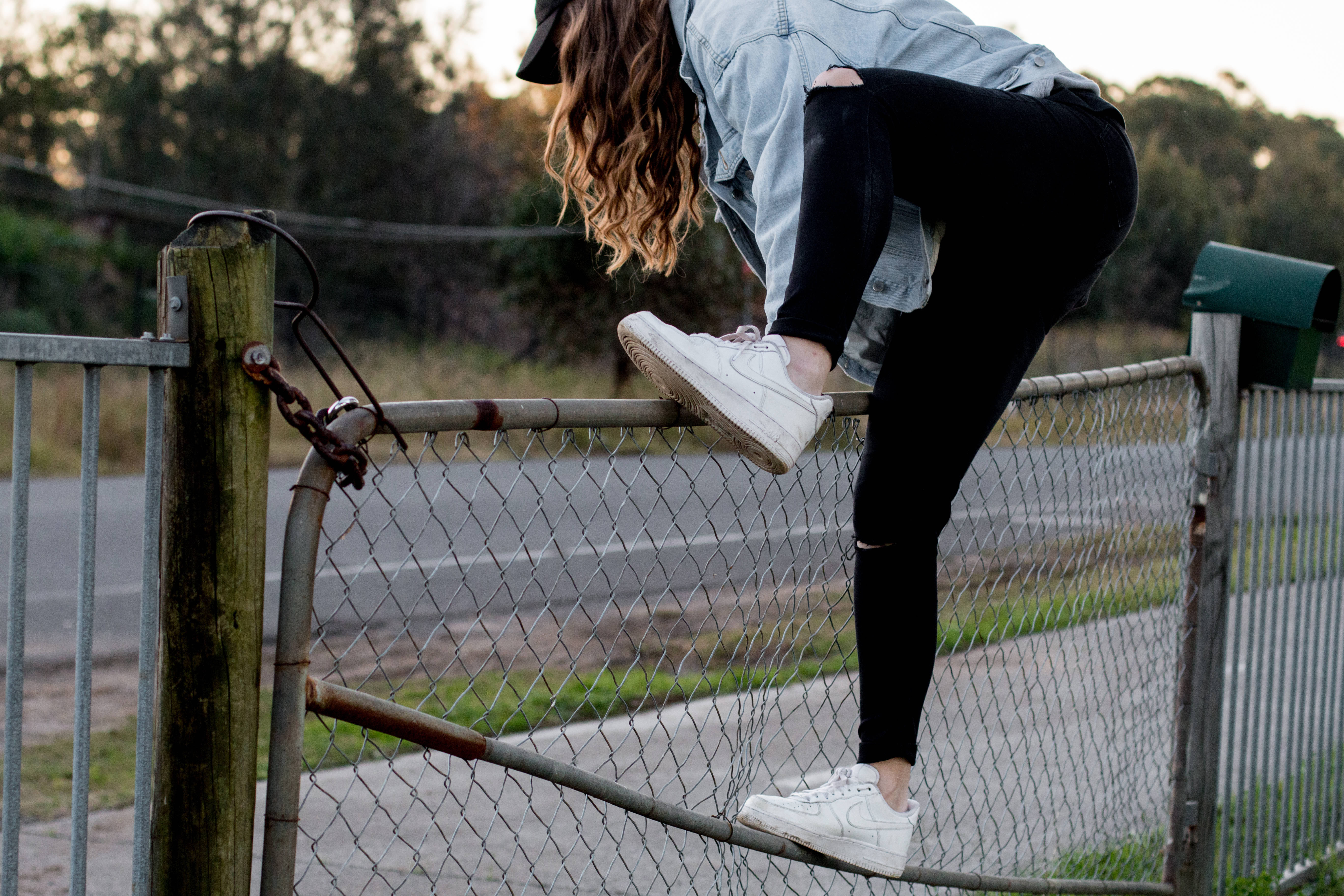 Parkour 桌布 壁紙 — 一位年輕女子穿著破洞牛仔褲和白色運動鞋，在黃昏時分動態地平衡在生鏽的鐵絲網圍欄上。
