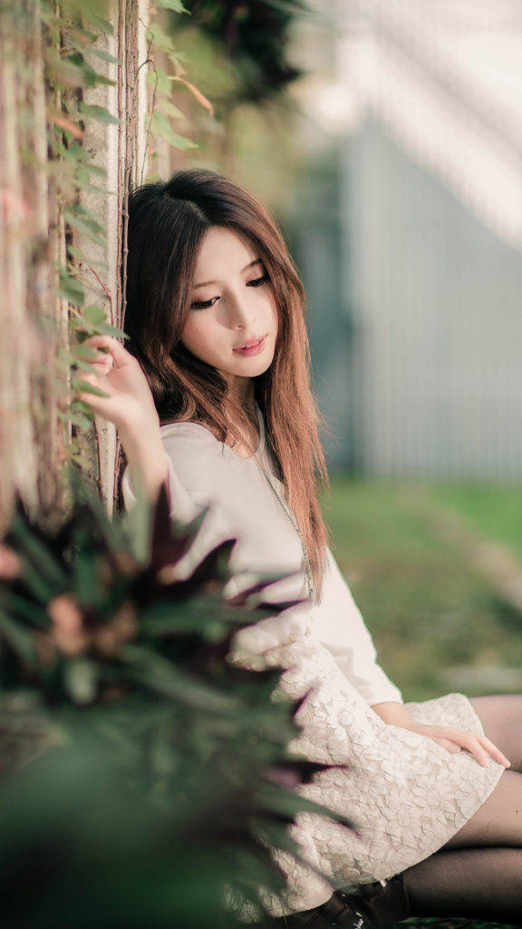 phone girl 桌布 壁紙 — 一位年輕女子身穿奶油色蕾絲連衣裙，在柔和自然光線下靠在一面爬滿藤蔓的牆上。