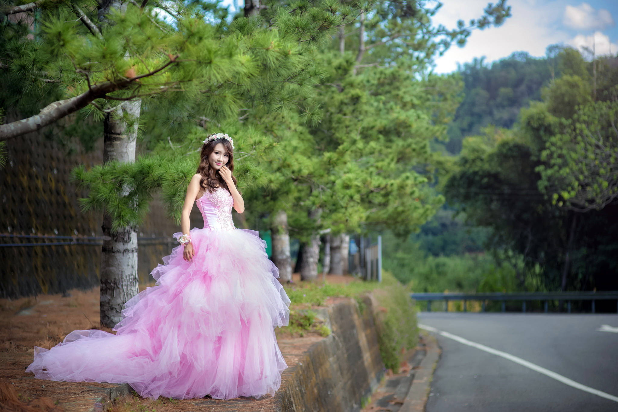 Bride 桌布 壁紙 — 一位年輕女子身穿寬大的粉紅色長裙，優雅地站在路邊的松樹旁，周圍是明亮的天空下鬱鬱蔥蔥的綠色森林。