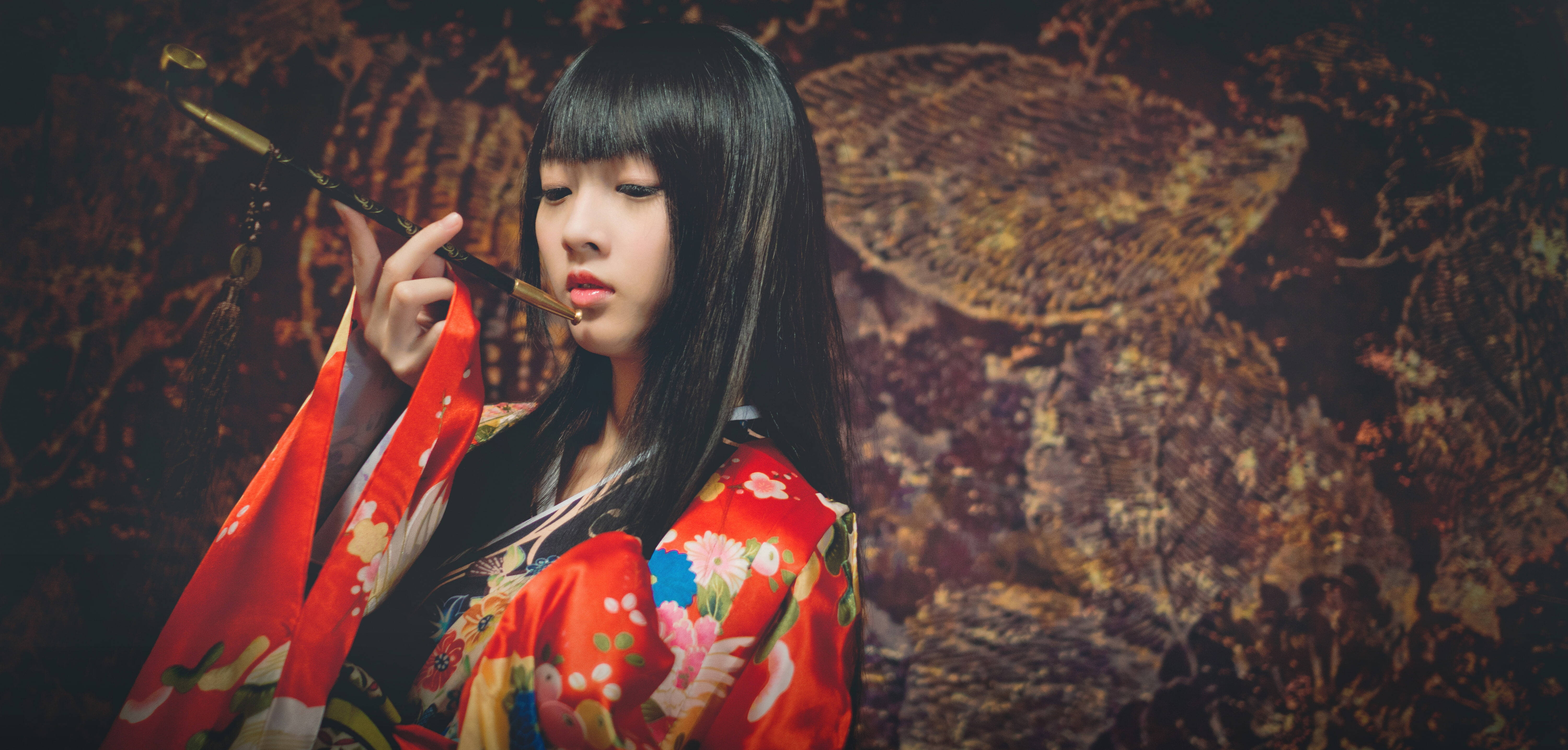 日本 桌布 壁紙 — 一位年輕女子身穿鮮豔的紅色花卉和服，手持傳統煙斗，背景為質感豐富的深棕色。