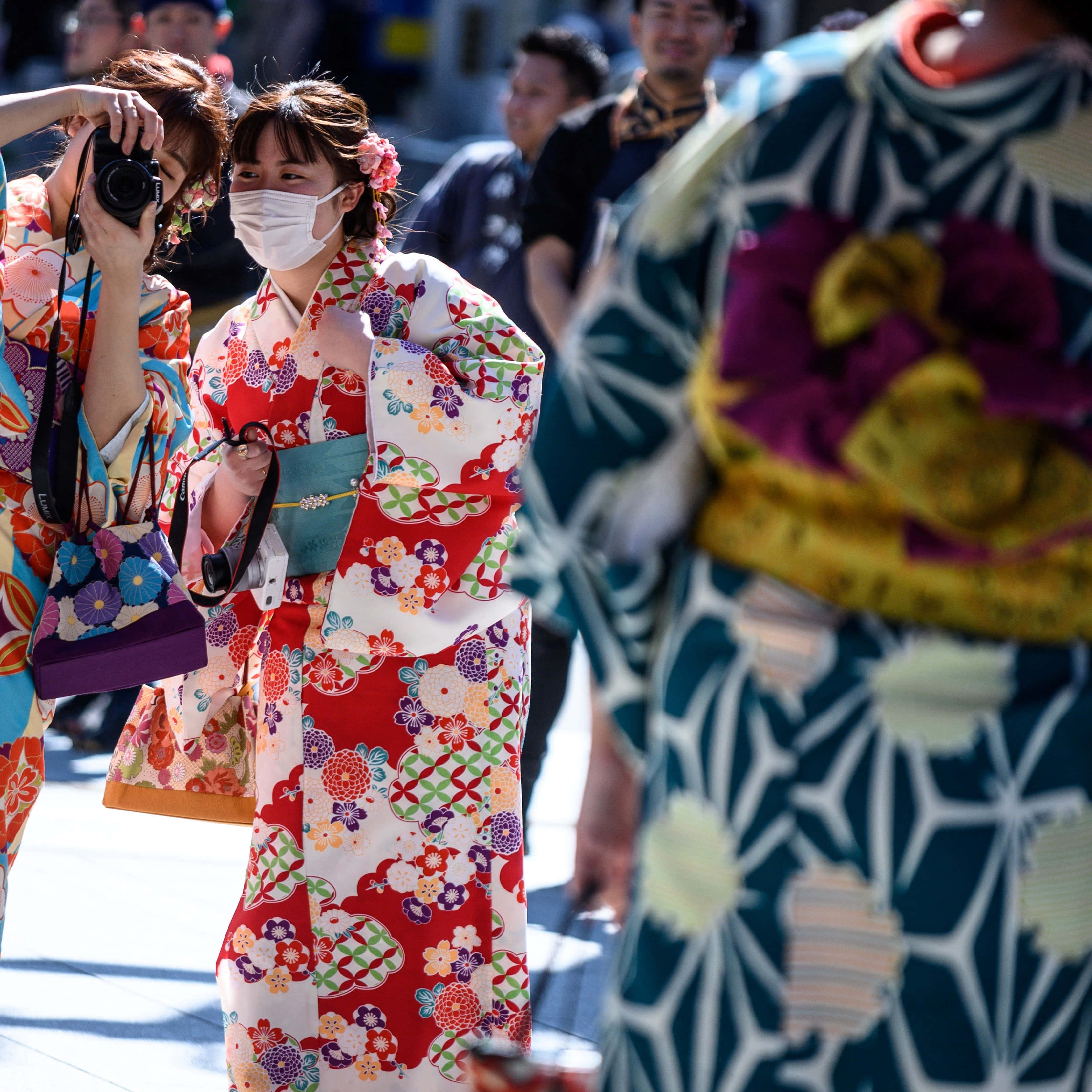Kimono 桌布 壁紙 — 一位年輕女子身穿鮮豔的紅色花卉和服，戴著白色口罩，走過繁忙的街道。