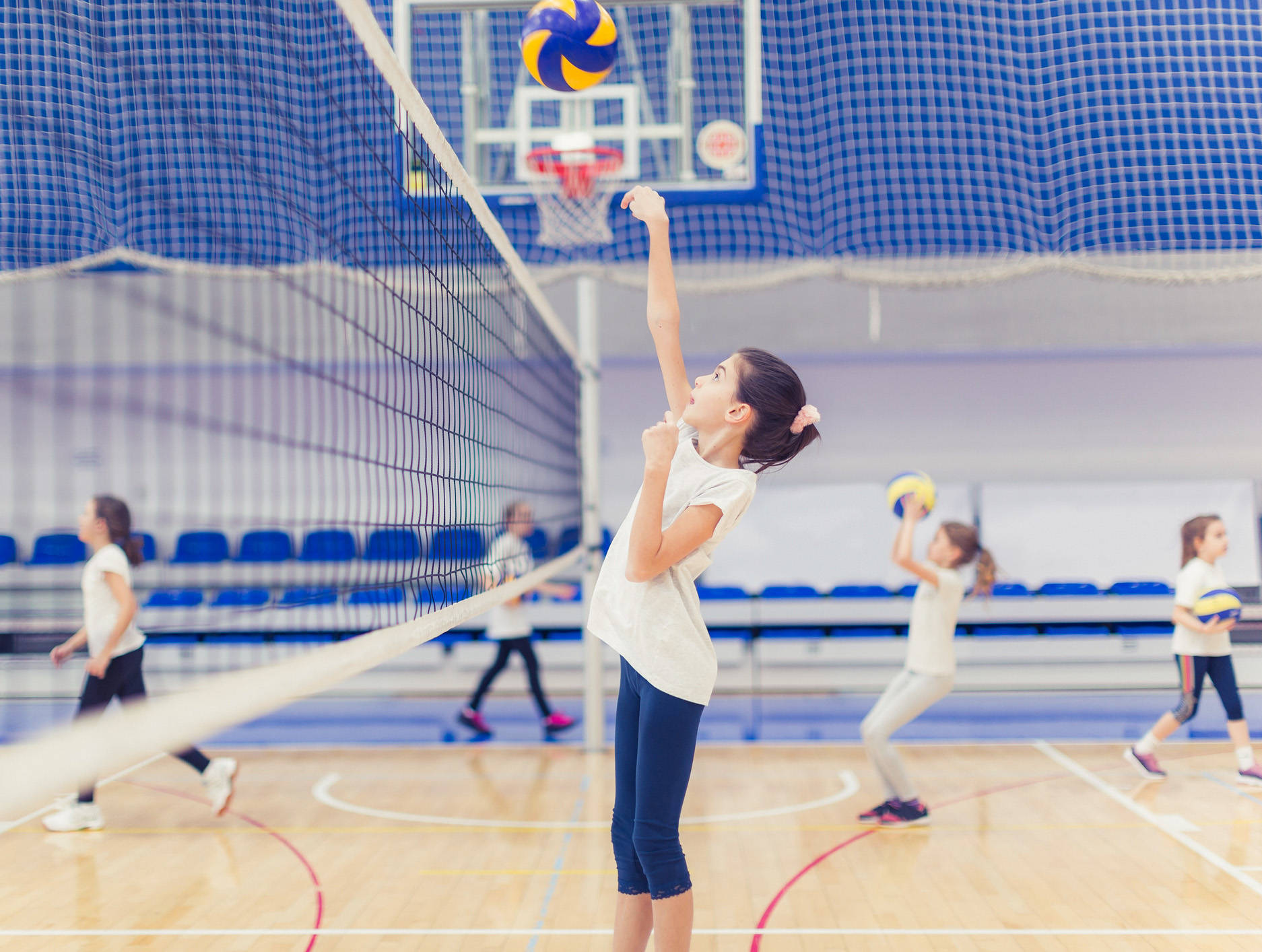 Volleyball 桌布 壁紙 — 一位年輕女孩在光線明亮的室內體育館跳起扣殺排球，背景有隊友在比賽。