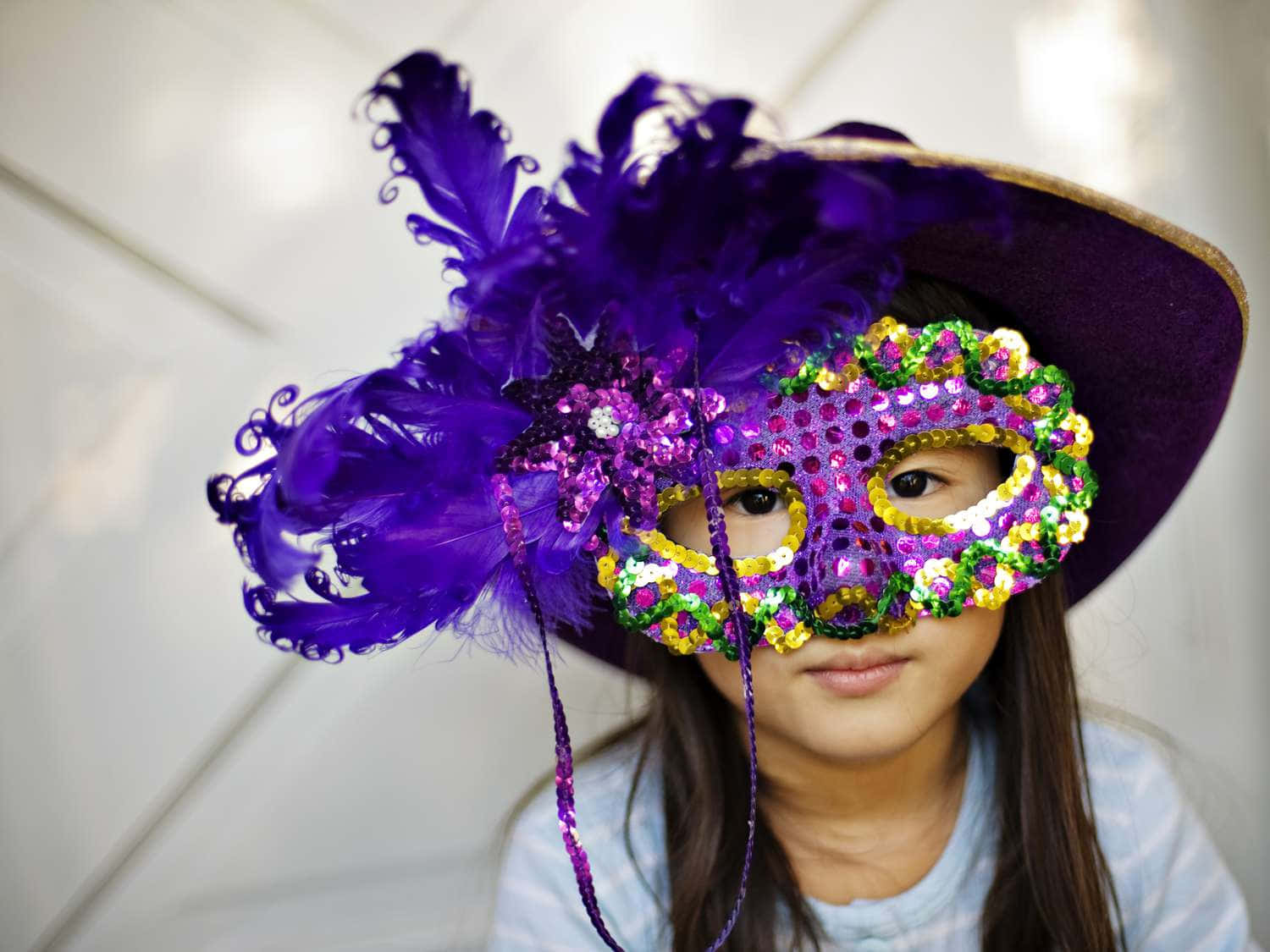 Mardi Gras 桌布 壁紙 — 一位年輕女孩戴著飾有亮片的鮮紫色羽毛面具和相配的帽子，背景是白色瓷磚牆。