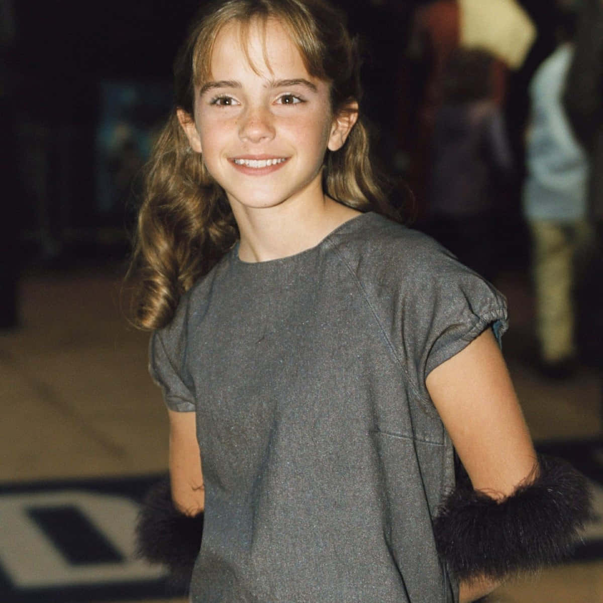 Emma Watson 桌布 壁紙 — 一位年輕女孩留著波浪般的棕色頭髮，穿著灰色連衣裙搭配黑色毛絨袖口，在溫暖的室內燈光下燦爛微笑。