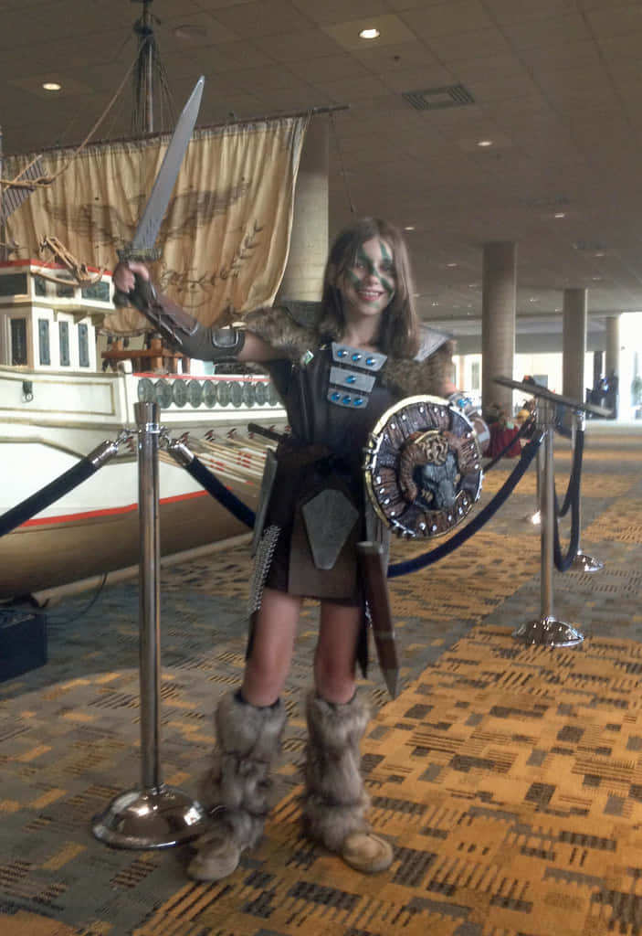 Aela The Huntress 桌布 壁紙 — 一位年輕女孩穿著精細的維京戰士服裝，在室內靠近船隻展覽處自豪地擺出持劍和盾牌的姿勢。