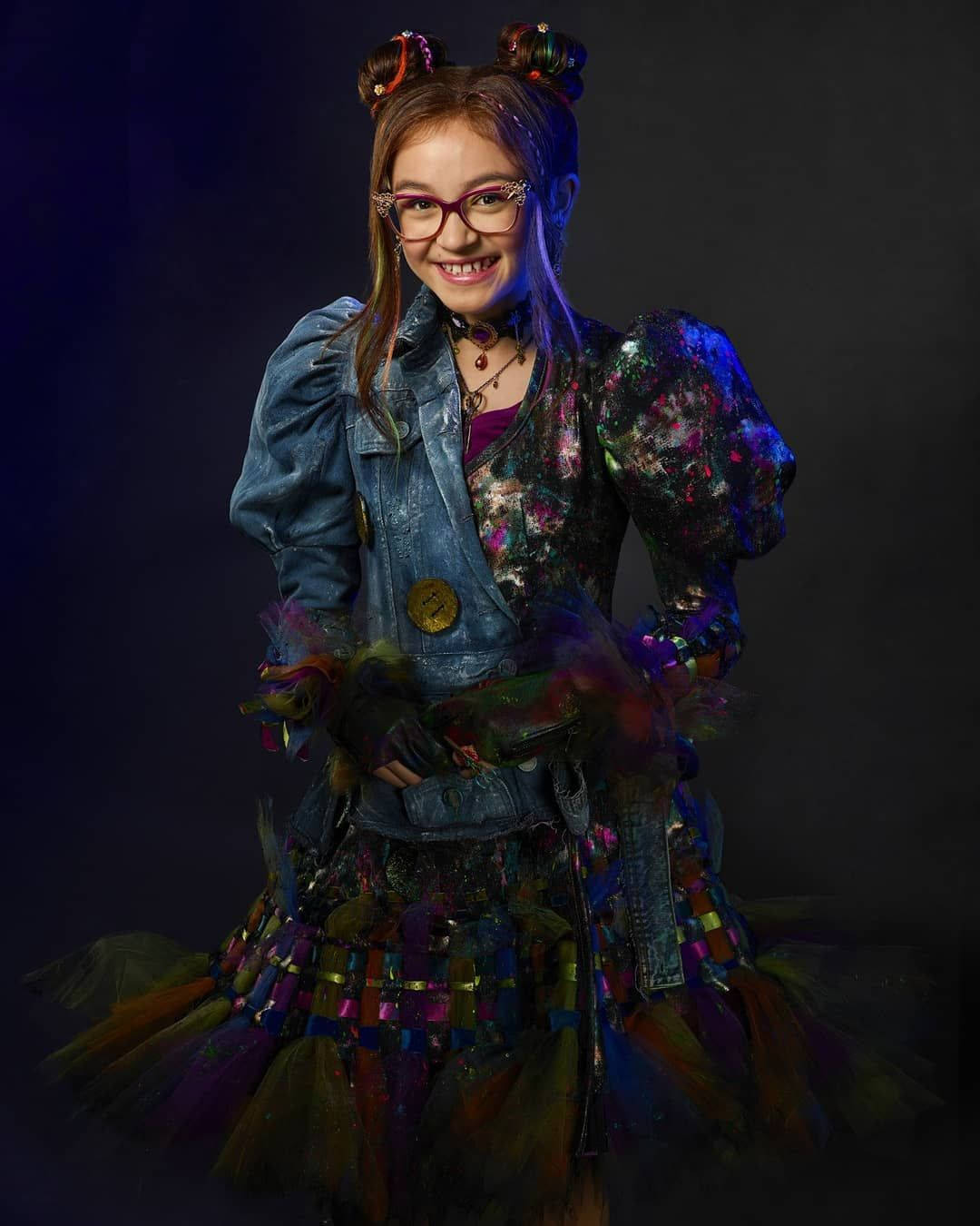 Descendants 桌布 壁紙 — 一位年輕女孩紮著彩色髮髻、戴著眼鏡，在深色工作室背景下穿著充滿活力、不拘一格的服裝，笑容燦爛。