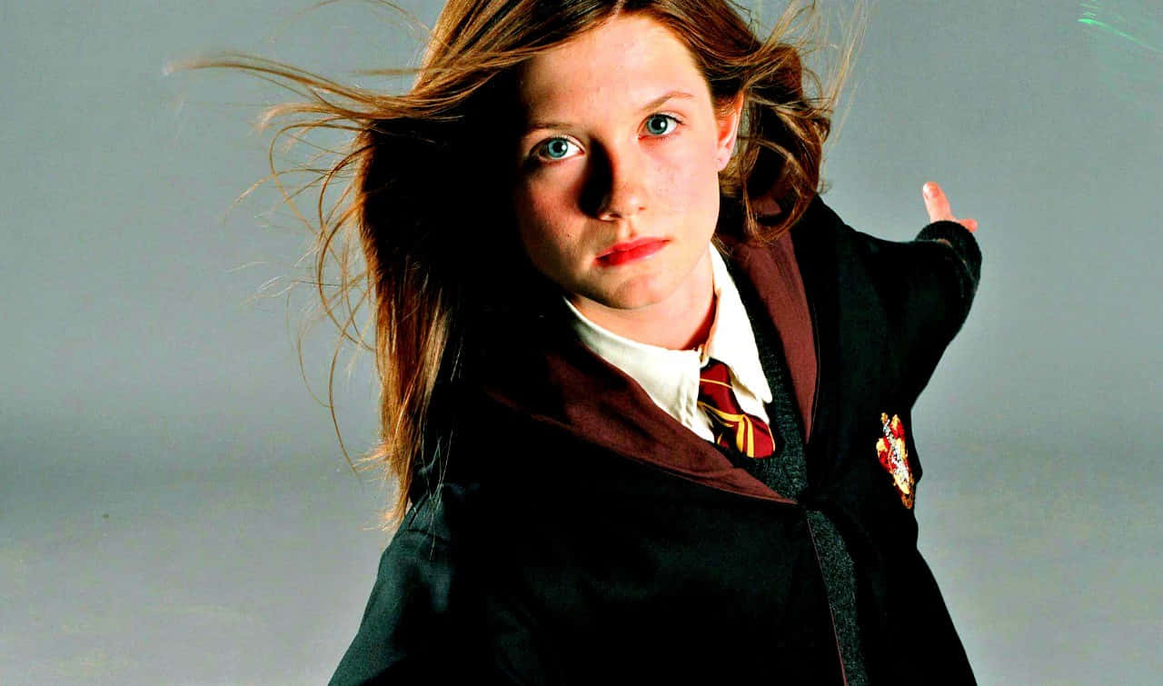 Ginny Weasley 桌布 壁紙 — 一位年輕女巫，頭髮被風吹起，眼神專注，身穿霍格華茲制服，背景是純灰色。