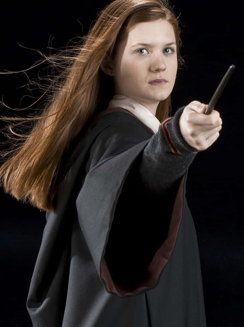 Ginny Weasley 桌布 壁紙 — 一位年輕女巫，飄逸的紅髮，在黑色背景前，戲劇性地在攝影棚肖像中向前指著魔杖。