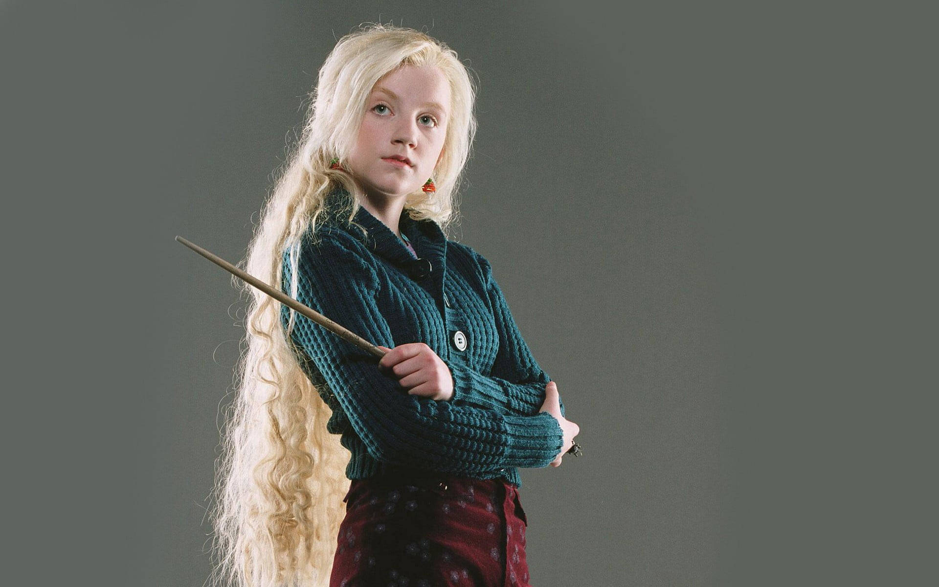 Luna Lovegood 桌布 壁紙 — 一位年輕女巫留著金色長髮，手持魔杖，身穿青綠色毛衣，背景為灰色。