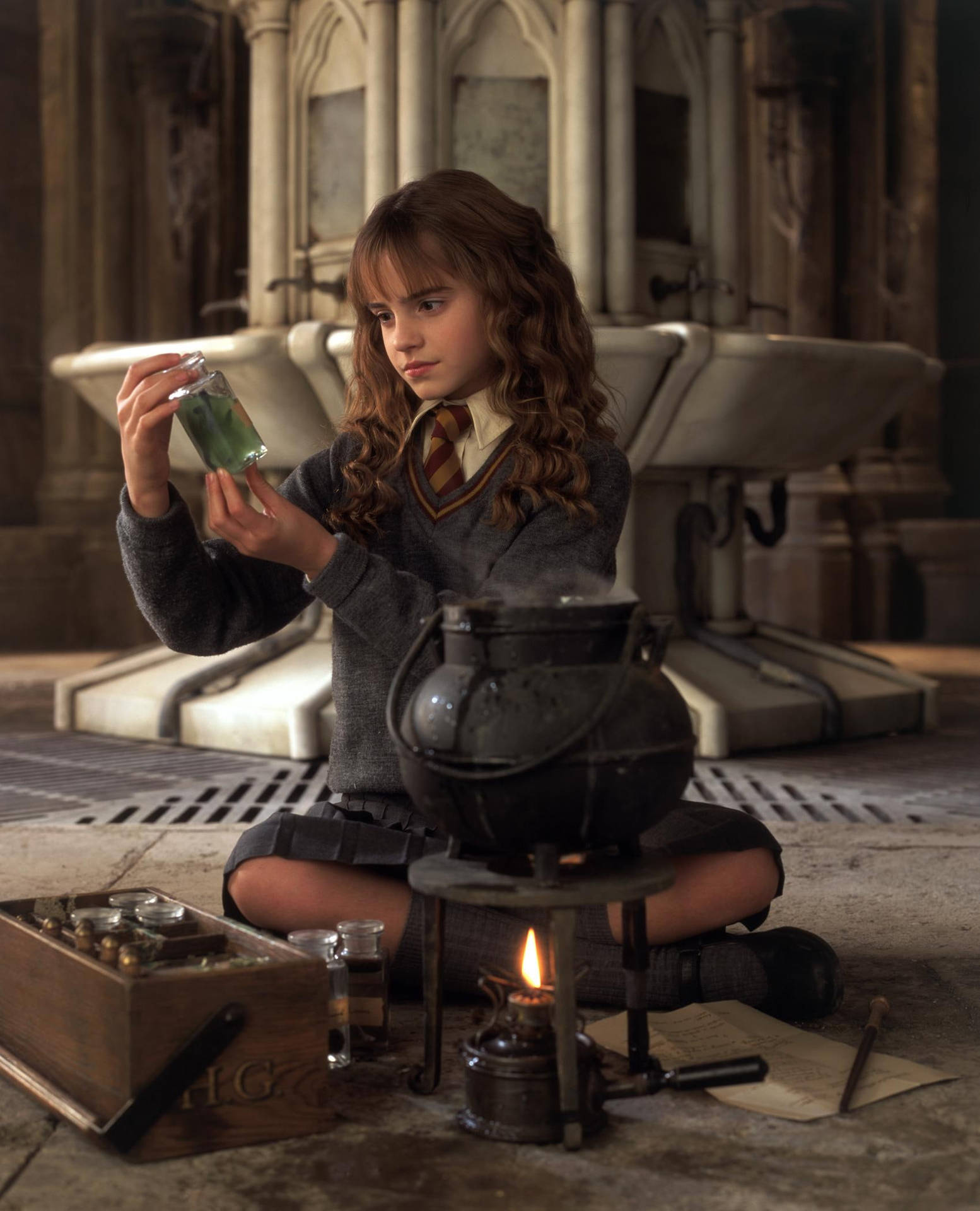 Hermione Granger 桌布 壁紙 — 一位年輕女巫跪在石頭地牢中，檢查一個發光的綠色藥水，旁邊是一個冒煙的大鍋。