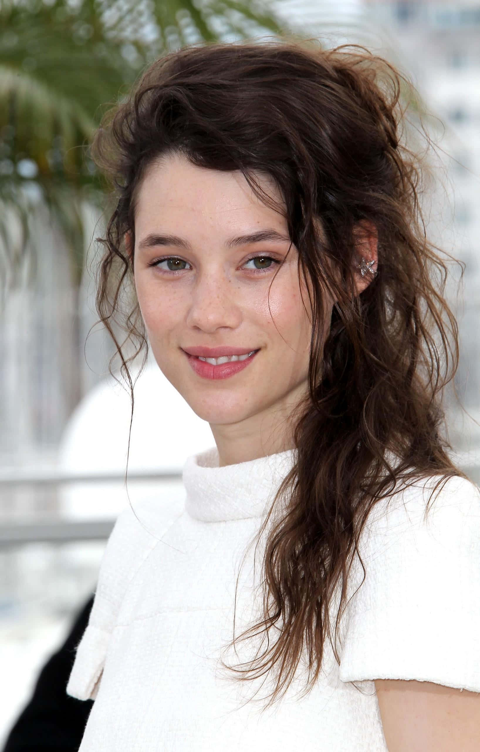Astrid Berges Frisbey 桌布 壁紙 — 一位年輕女性，一頭凌亂的棕色頭髮，帶著溫柔的微笑，身穿白色質感上衣，背景是模糊的棕櫚樹。
