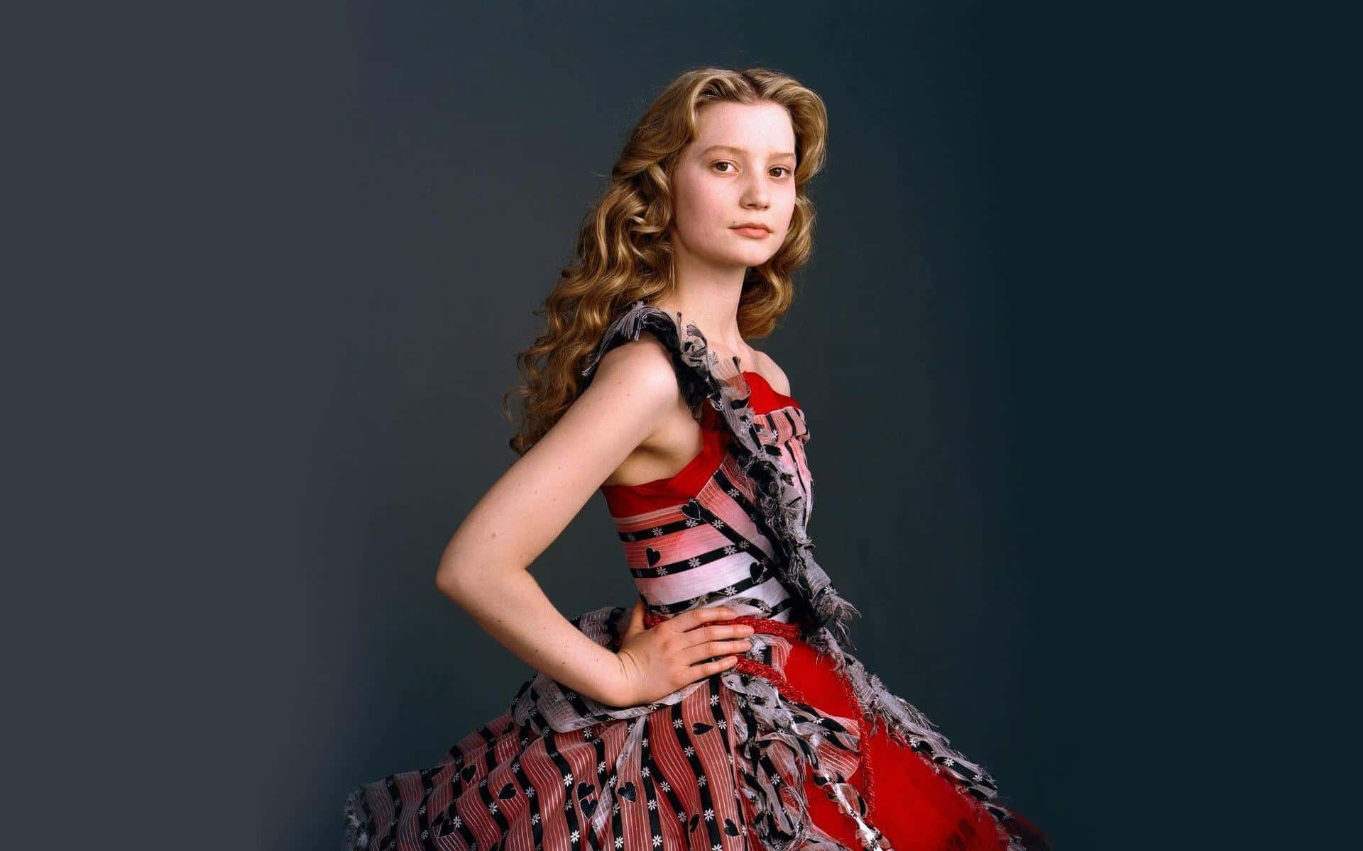 Mia Wasikowska 桌布 壁紙 — 一位年輕女性，一頭捲曲金髮，身穿紅黑圖案連衣裙，在深灰色攝影棚背景前自信地擺姿勢。