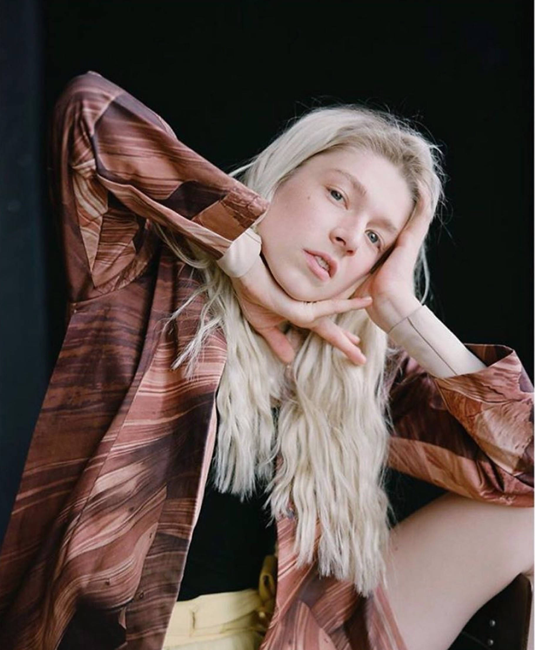 Hunter Schafer 桌布 壁紙 — 一位年輕女性，一頭鉑金色頭髮，在純黑背景前將下巴擱在手上，身穿飄逸的棕色圖案襯衫。