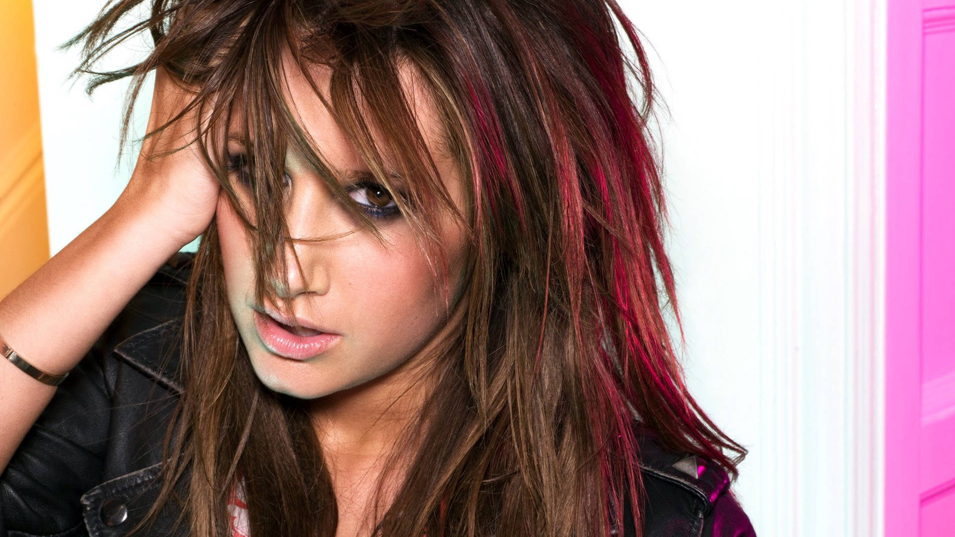 Ashley Tisdale 桌布 壁紙 — 一位年輕女性，凌亂的棕色頭髮帶有紅色條紋，將頭靠在手上，靠著一面亮粉紅色和黃色的牆。