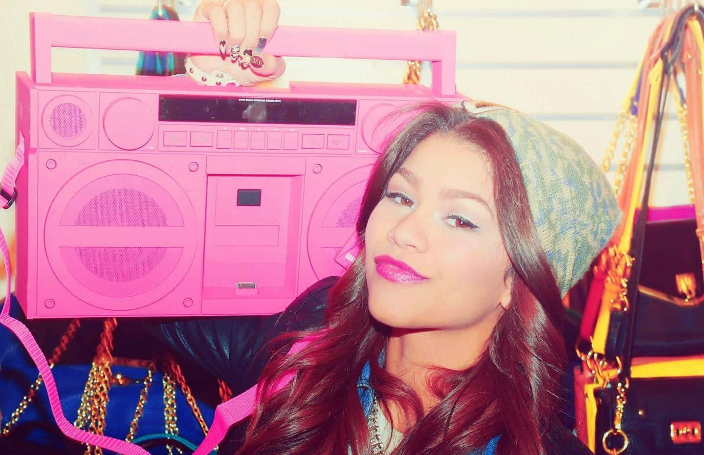 Zendaya 桌布 壁紙 — 一位年輕女性，化著大膽的妝容，戴著圖案無邊帽，自信地站在一個巨大的粉紅色音箱旁，音箱上有色彩繽紛的配件。