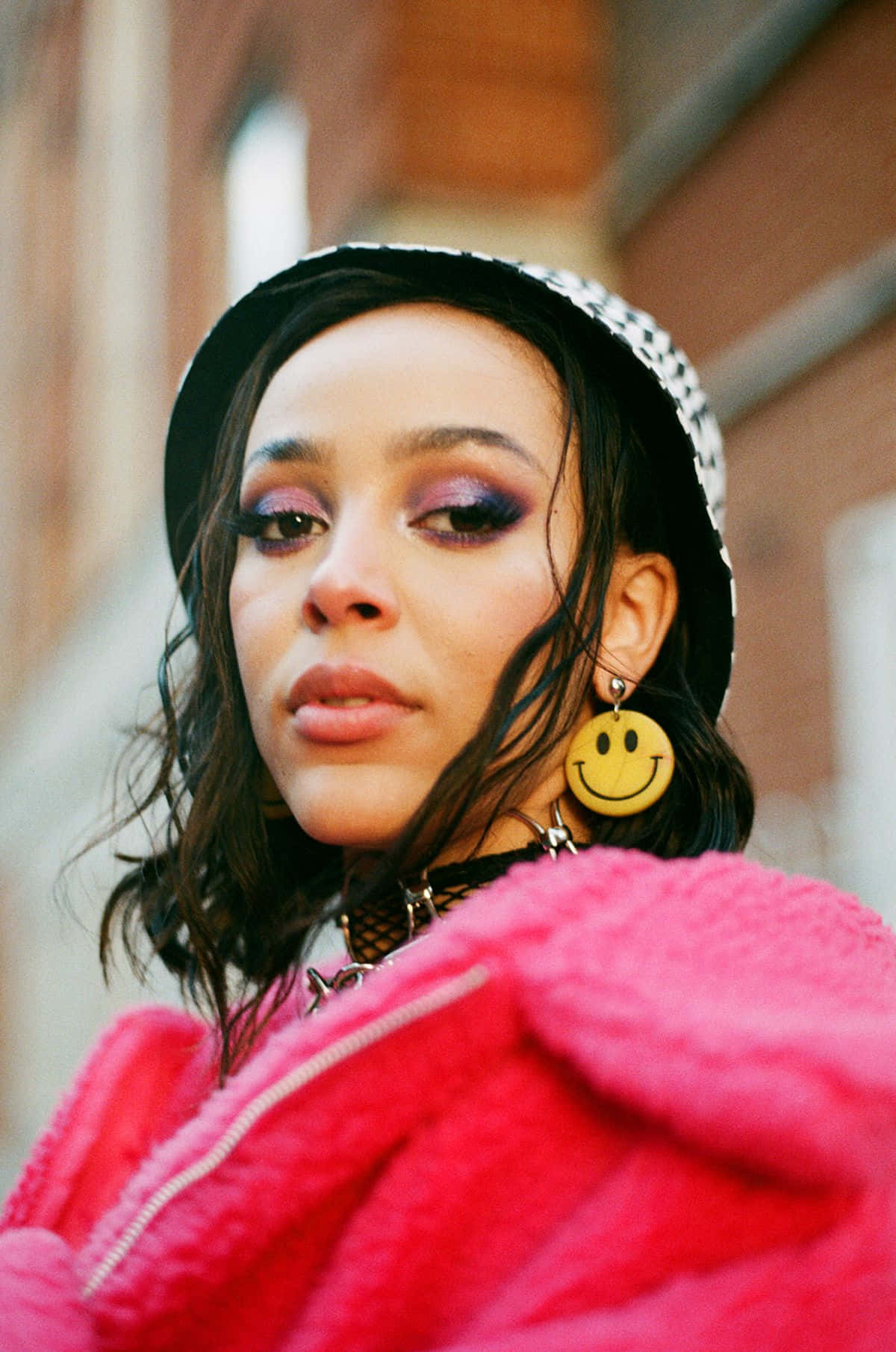 Doja Cat 桌布 壁紙 — 一位年輕女性，化著大膽的紫色眼妝，穿著亮粉色的毛絨外套，戴著黑白格紋帽子和黃色笑臉大耳環，直視鏡頭。