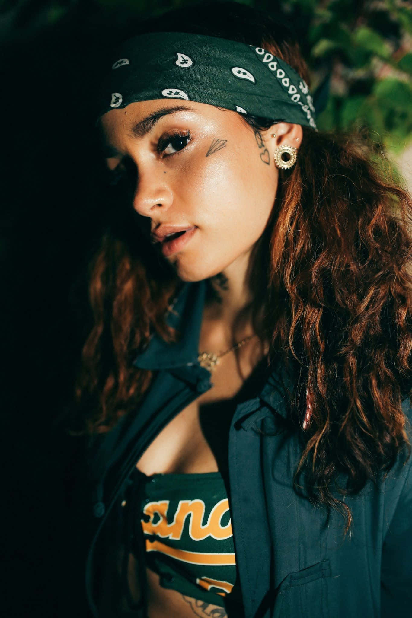 kehlani 桌布 壁紙 — 一位年輕女性，捲髮戴頭巾，在昏暗、樹葉茂密的環境中若有所思地凝視。