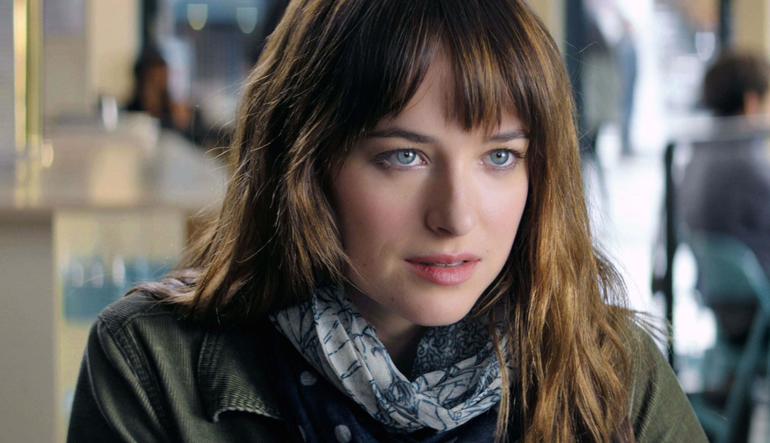 Dakota Johnson 桌布 壁紙 — 一位年輕女性，擁有引人注目的藍色眼睛和棕色頭髮，戴著圖案圍巾和綠色外套，若有所思地向前凝視。
