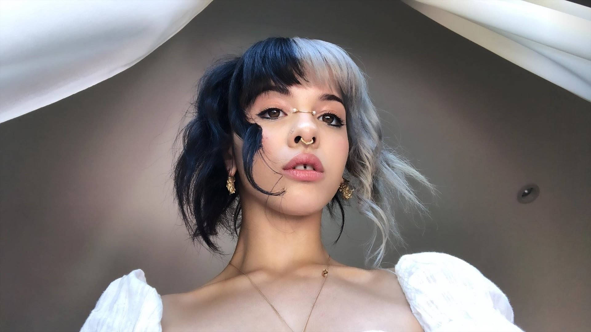 Melanie 桌布 壁紙 — 一位年輕女性，擁有引人注目的黑銀色頭髮和金色珠寶，在垂墜的白色布料下自信地擺姿勢。