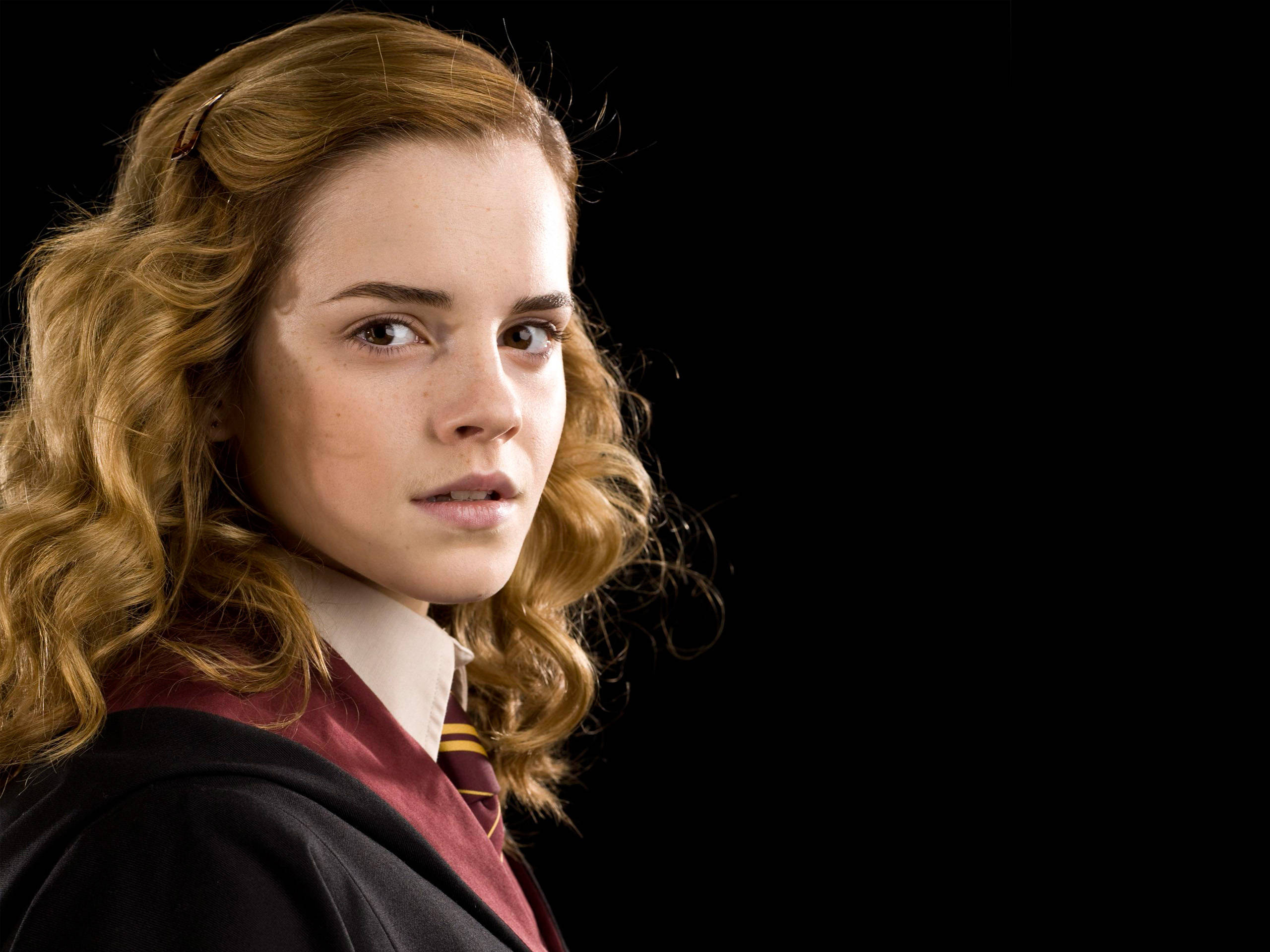 Hermione 桌布 壁紙 — 一位年輕女性，擁有捲曲紅髮，穿著栗色和金色的校服，背景為純黑色。